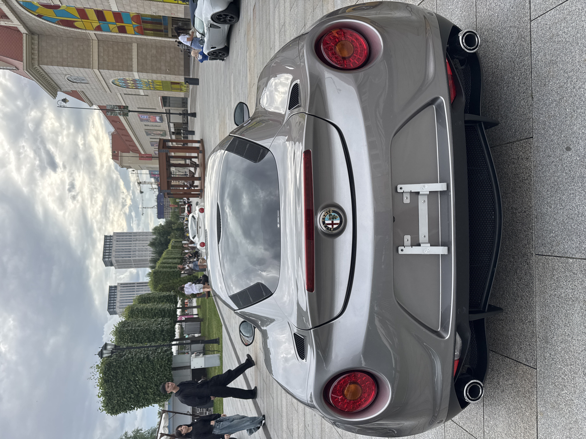 The image captures a rear view of a grey Alfa Romeo 4C sports car parked on a paved pedestrian walkway in a modern urban environment. The car's brake lights and taillights are illuminated. In the background, several people are walking and standing. To the left, two individuals, a woman and a man in a black outfit, are walking away from the camera. Further back, a line of manicured, box-shaped trees separates the walkway from a grassy area with streetlights. Beyond the trees, two tall, modern buildings with light-colored facades and dark windows dominate the skyline. To the right of the car, a portion of a building with a red roof and a large, colorful stained-glass window is visible, suggesting a commercial or entertainment complex. Another white sports car is parked in the background on the right, with a woman and a child near it. The sky is overcast, with scattered clouds. The scene appears to be a public space in a city, possibly a promenade or a plaza. The Alfa Romeo 4C is the primary subject, positioned centrally in the frame. The overall impression is one of a car show or a leisure event in a city. This scene is located at the Moscow City complex, specifically near the "Afimall City" shopping center.