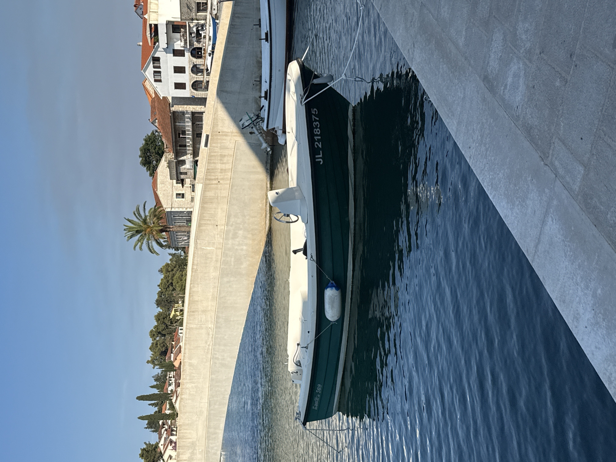 A small, dark green and white motorboat with the registration "JL 218375" is moored in the clear blue water of a harbor in Jelsa, Croatia. The boat is tied with ropes to a white mooring buoy and the shore. In the background, a concrete bridge spans across the water, casting a shadow on the boat. Beyond the bridge, a row of buildings with red-tiled roofs and white facades line the waterfront, interspersed with trees and a palm tree. The sky is clear and blue, with a hint of clouds. The scene appears to be a quiet, sunny day in a coastal town.
