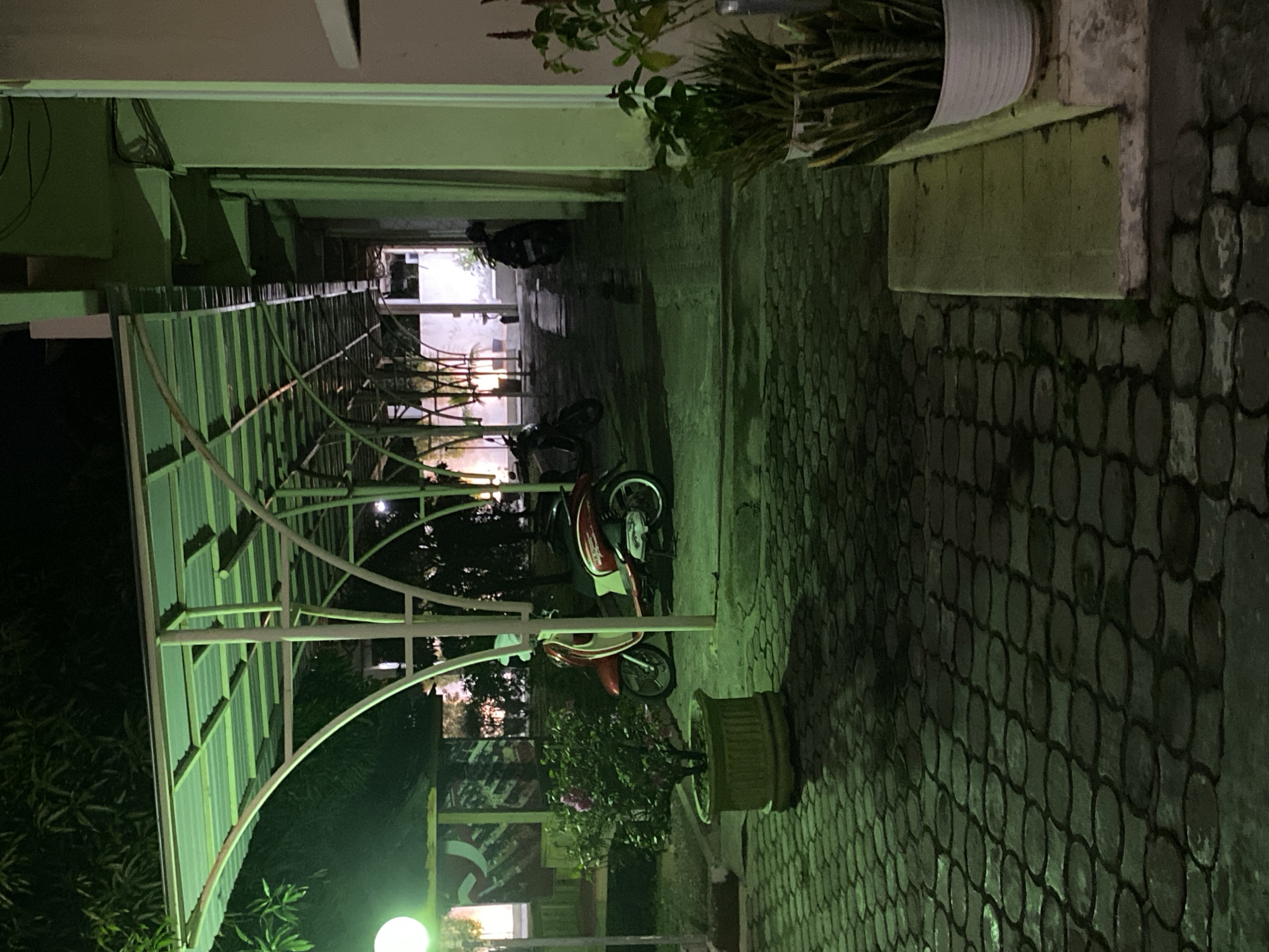 An exterior, night-time shot depicts a covered walkway with a green-tinted metal roof supported by white metal pillars. Paving stones form the ground surface, which appears wet in places, reflecting the green light. A potted plant with pink flowers sits to the left. Two red and white scooters are parked under the walkway. Further down the walkway, more scooters and plants are visible. The scene is illuminated by artificial green lighting from the walkway's roof and a bright white circular light source to the left of the frame. The background shows dark foliage and a building with a light-colored wall. The overall impression is one of a well-lit outdoor space at night, likely a parking area or pedestrian pathway.