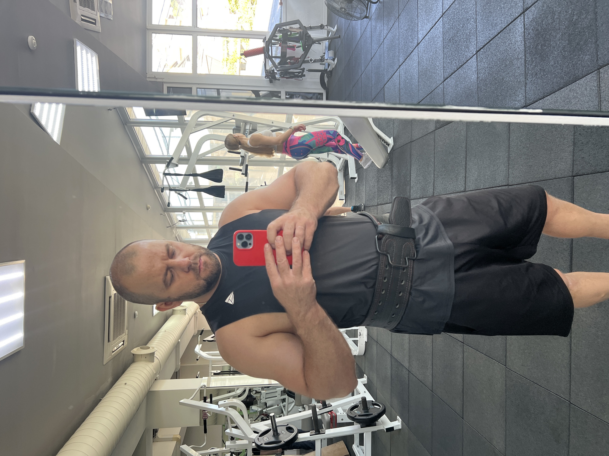 This image is a mirror selfie taken by a man in a modern gym, likely located in Sochi, Russia.
The primary subject is a man, seen from the chest down to his upper thighs, taking the photo with a red smartphone held in his right hand. He has a shaven head and a short beard, with his eyes closed as the photo is taken. He is wearing a black sleeveless tank top featuring a white, stylized triangular or arrow-like logo on the chest, black shorts, and a dark, sturdy weightlifting belt around his waist. His left arm is bent, showcasing muscular development.
Reflected in the background is a woman with long, blonde hair, appearing to lie or rest on a white gym bench. She is dressed in a white sports bra and brightly colored leggings with a geometric pattern in shades of pink, blue, and green. The text "MARCY" is visible on the side of the bench she is using.
The gym environment is bright, with large windows along one wall allowing significant natural light, indicating it is daytime. Green foliage is visible outside the windows. The interior features a light gray wall, a dark gray tiled floor, and a ceiling with exposed white ductwork, rectangular LED light panels, and a wall-mounted air conditioning unit. Various pieces of gym equipment, including weight racks, benches, and loose weights, are scattered throughout the space. A black industrial fan is also visible in the upper right corner of the reflection.
The overall scene depicts a functional and well-lit fitness facility where individuals are engaged in or resting from their workouts. The man's attire and the weightlifting belt suggest an intention for heavy lifting.