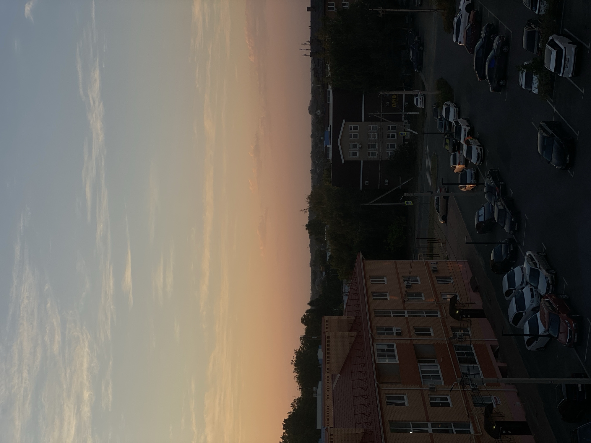 The image shows an aerial view of a residential area at dusk in Zelenopol'skii, Russia. The sky is transitioning from a pale blue at the top to an orangey-pink hue near the horizon, with wispy clouds scattered across it. In the foreground, a light-orange brick apartment building with a red tiled roof is visible. The building has multiple windows and balconies. Adjacent to it is a parking lot filled with various cars. Further in the background, more buildings, trees, and a distant treeline are observable. The overall scene is quiet and depicts a typical urban or suburban environment during the evening hours. There are no people visible in the image.
