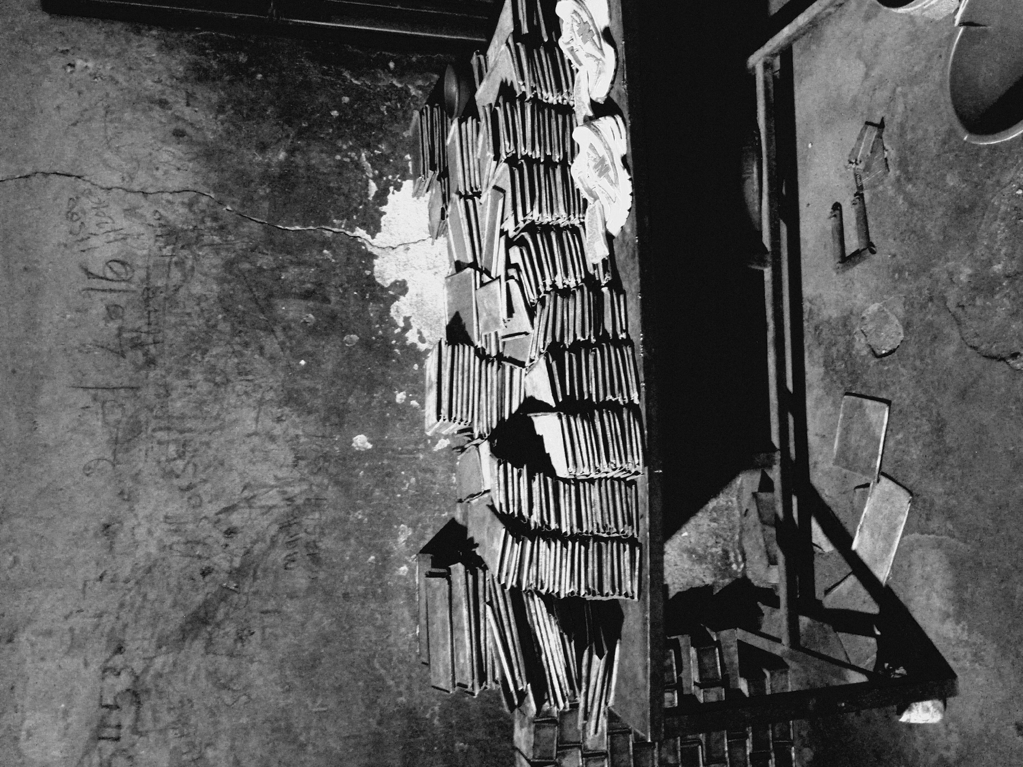 The image shows a room in Unknown, Unknown. The room has a concrete floor with cracks and graffiti. In the center of the room is a stack of rectangular metal plates. There is a shoe on top of the metal plates. Adjacent to the stack of plates is a metal structure. The image is in black and white.