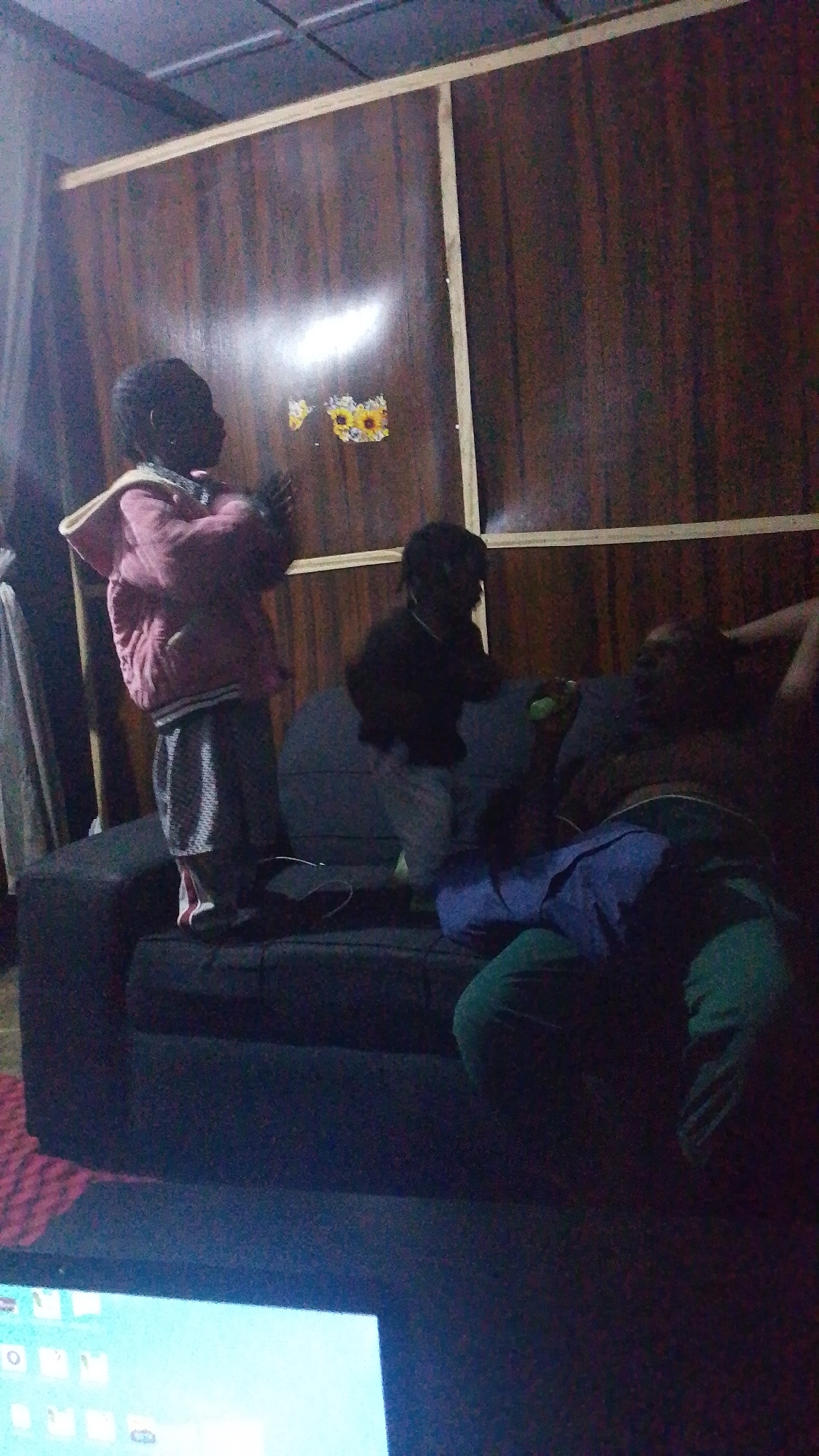 Three people, two children and one adult, are on a dark grey couch in a dimly lit room. The child on the left is wearing a pink jacket and striped shorts, standing on the couch with hands raised. The child in the middle is wearing dark clothing and light-colored pants, sitting on the couch. The adult on the right is lying on the couch with a blue shirt and green pants. A light-colored object, possibly a bottle or toy, is in the adult's hand. The background features a wooden paneled wall with a decal of sunflowers. A portion of a laptop screen is visible in the foreground. The room appears to be indoors, possibly a living room, in an unknown location.