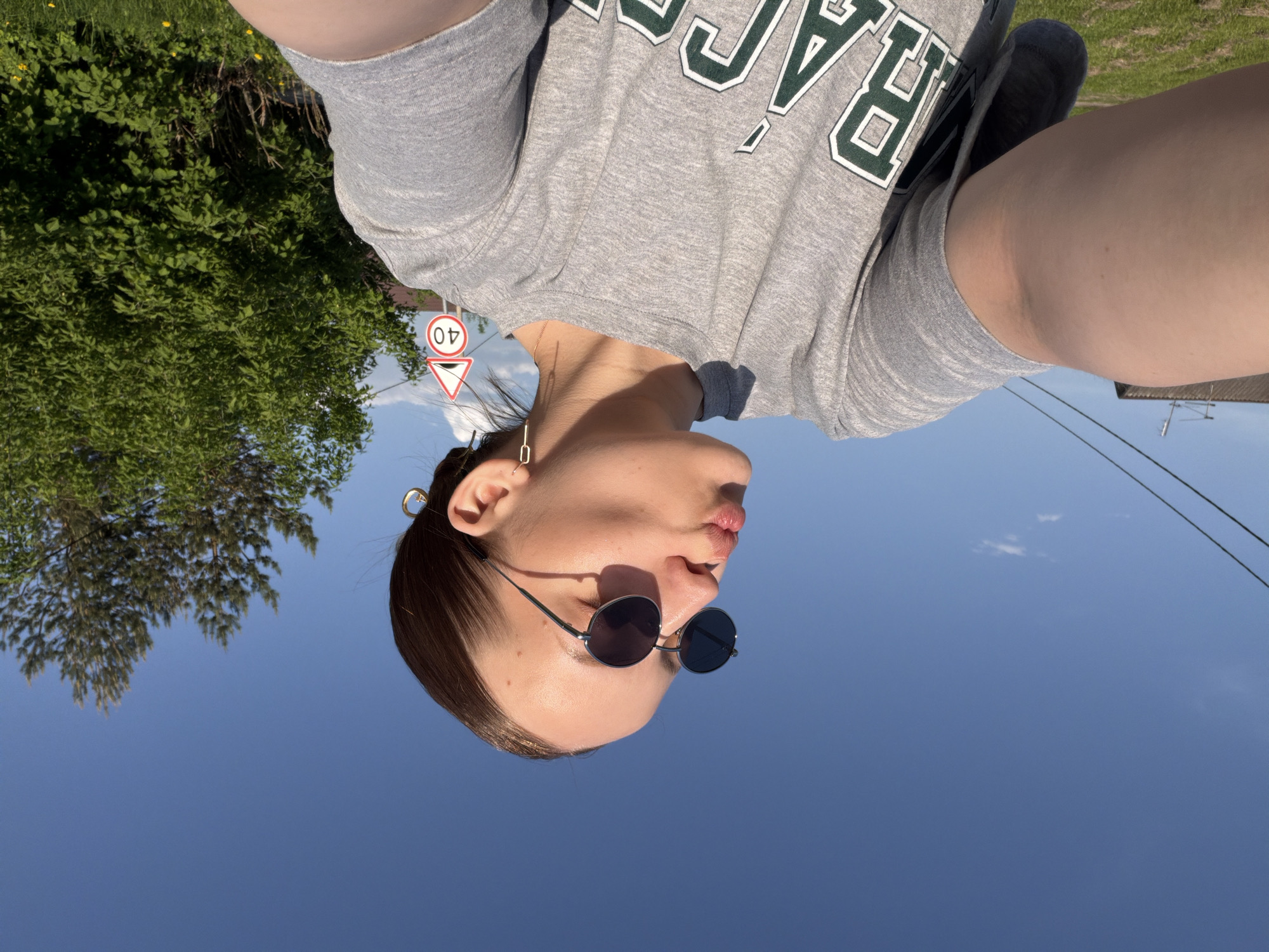 A low-angle, upside-down shot shows a woman wearing round sunglasses and a gray t-shirt with a logo. She is outside on a sunny day with a clear blue sky. A leafy green tree is on the left side of the frame and power lines are on the right. A speed limit sign is partially visible behind the woman’s neck. This image was most likely taken in the city of Reshetnikovo, Russia.