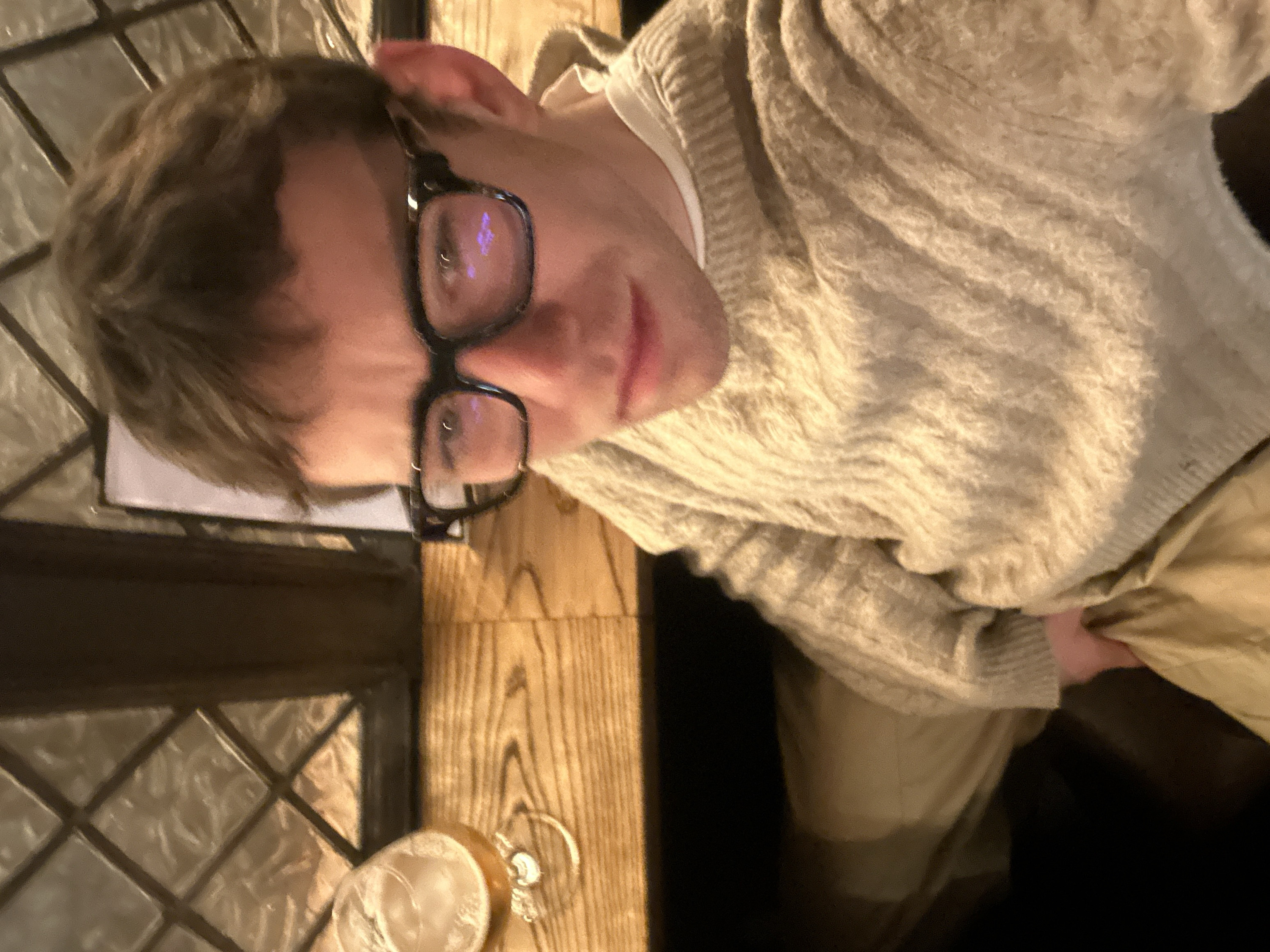 A person with short, light brown hair and black-framed glasses looks toward the camera. They are wearing a cream-colored knit sweater over a white shirt. The individual sits at a wooden table next to a leaded glass window. A glass of liquid rests on the table beside a small, gold-colored object, possibly a bottle opener. The person's pants appear to be tan. The overall scene has a warm, indoor ambiance.