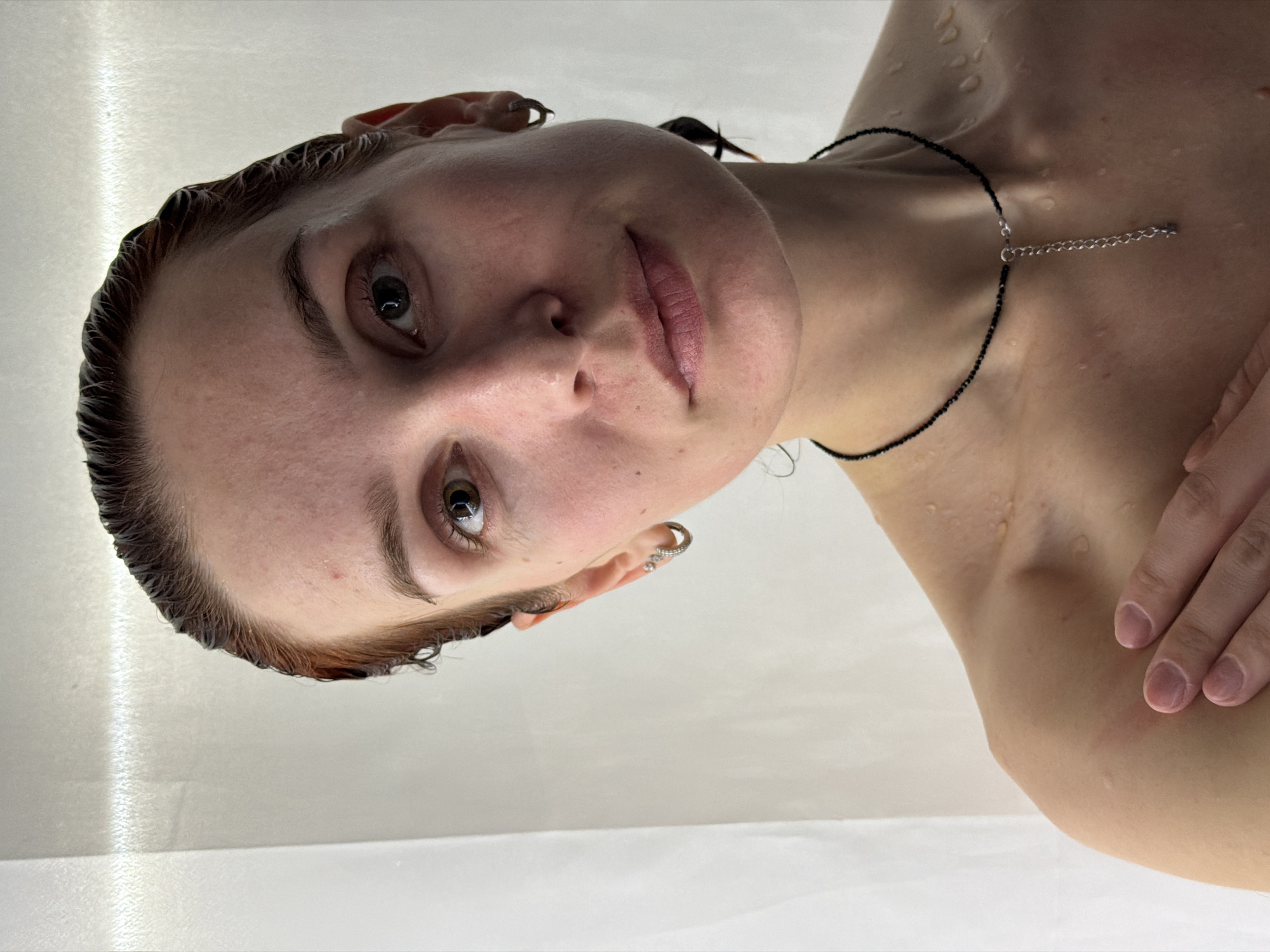 A fair-skinned individual with damp, short brown hair is shown from a low-angle perspective. Water droplets are visible on the face, neck, and upper chest. The person has light eyes, full lips, and pierced ears with jewelry. They wear a black beaded necklace with a silver extension chain. The background is a plain, light-colored surface, possibly a shower or bathroom wall. The person's right hand is partially visible, resting near their shoulder. The image's origin in Chaikovsky, Russia, is not discernible from the visual content.