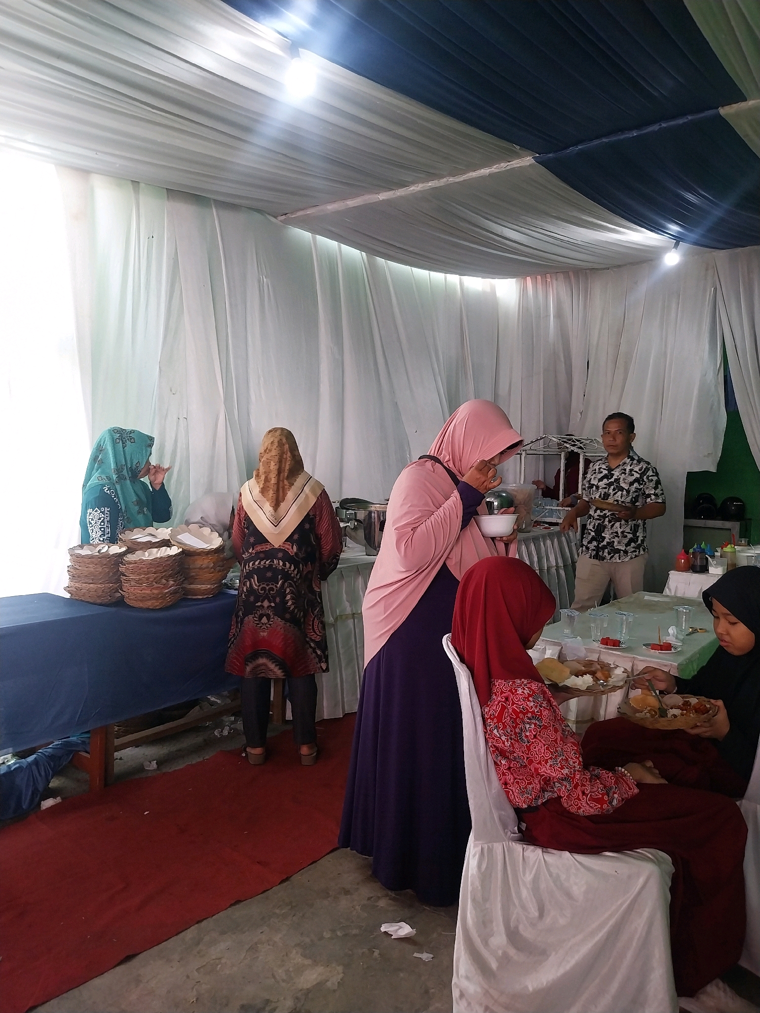The image depicts a gathering, possibly a wedding reception or catered event, set up under a tent in Cirebon, Indonesia. White and navy fabric drapes the ceiling, with bright lights illuminating the space. Several individuals are present, some attending to food service and others seated at tables. In the foreground, two young females are seated at a table, one wearing a red hijab and patterned top, the other a black hijab and dark clothing. They are engaged with plates of food. To the right, a woman in a pink hijab and purple dress is holding a bowl and appears to be eating. Further back, a man in a patterned shirt and khaki pants stands behind a buffet table, holding a plate. Two women are visible near stacks of disposable plates on a table covered with a dark blue cloth. One, wearing a teal hijab, is interacting with the other, who has a brown and beige patterned hijab and a colorful dress. The environment suggests an outdoor or semi-open event space, with a red carpet runner on the ground. The overall scene suggests a social gathering where food is being served and consumed.