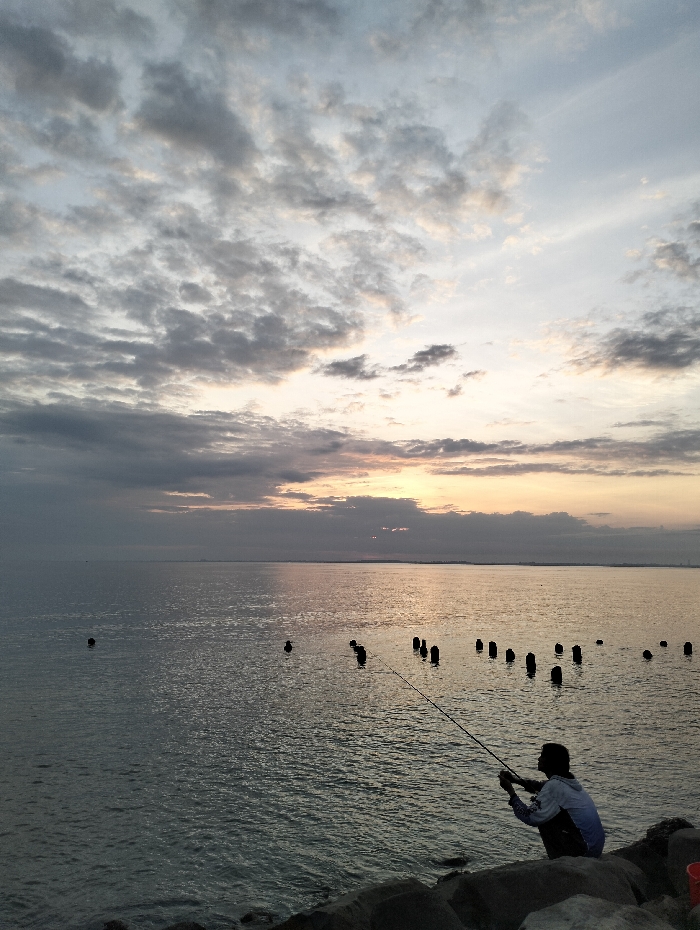 A person is fishing on rocks by a body of water. The scene features a sky filled with clouds, suggesting either dawn or dusk. Several pilings are visible in the water, adding depth to the view. The overall composition conveys a sense of tranquility, with the solitary figure engaged in the act of fishing. The lighting is muted, creating a soft and atmospheric effect.