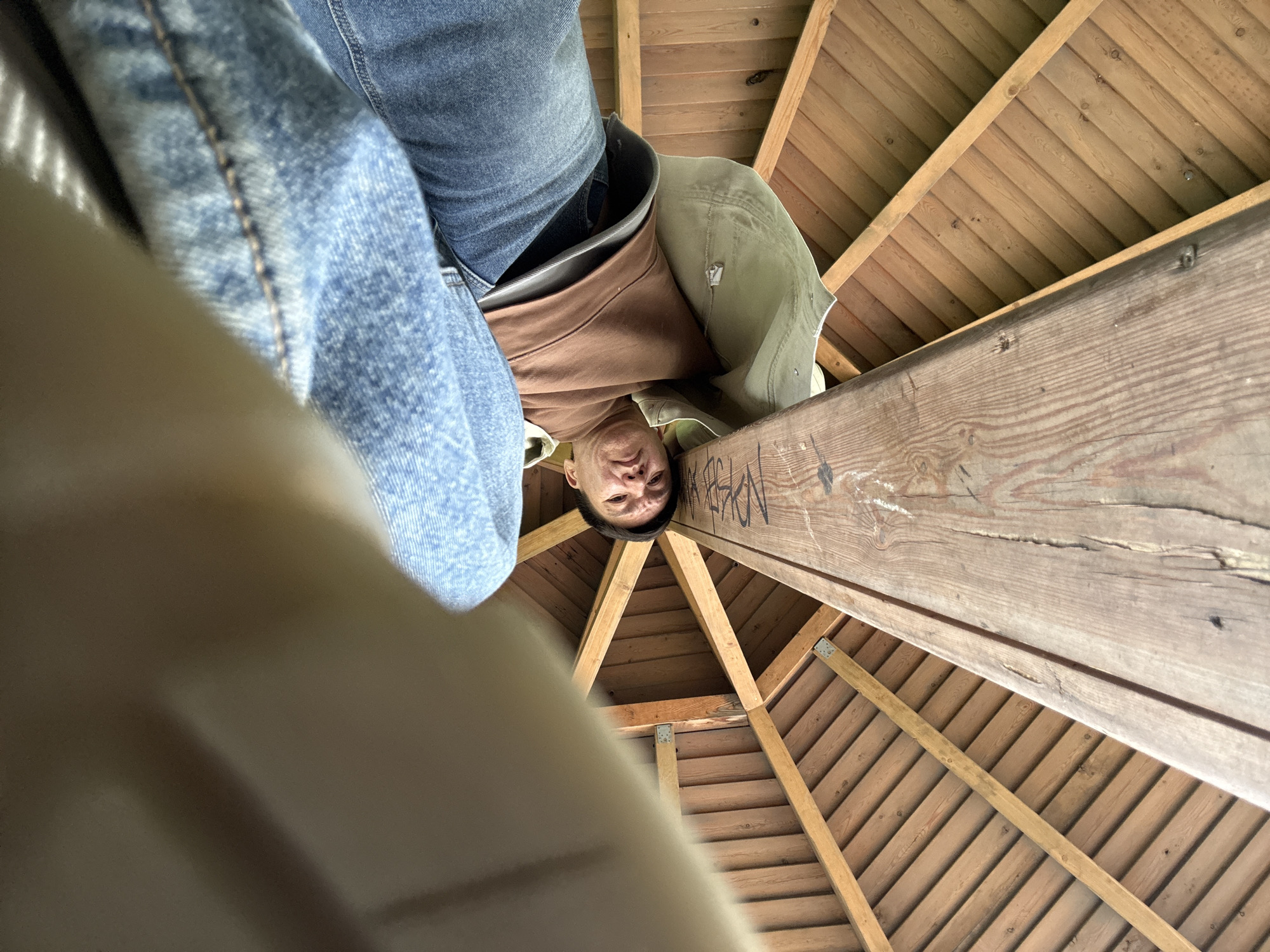 This low-angle, wide-perspective photograph looks upward into a covered wooden structure. A man's face, positioned upside down relative to the camera's orientation, is visible in the upper-middle of the frame, looking directly down at the lens with a neutral or slightly strained expression. He is dressed in blue jeans, a brown long-sleeved shirt, and a light olive green jacket.
The setting is an interior or underside of a wooden ceiling, likely part of a gazebo, lookout tower, or similar outdoor pavilion, typical of a park or recreational area in Moscow, Russia. The ceiling is constructed from numerous light-colored wooden planks arranged in a radial pattern, supported by sturdy wooden beams that converge towards a central point.
A blurred, light-colored object, possibly clothing or a surface, occupies the bottom-left foreground, close to the camera lens. The lighting suggests daytime, likely overcast or diffused, as there are no harsh shadows.
Graffiti is visible on a prominent, weathered wooden beam positioned to the right of the man's head. The text is partially decipherable, appearing to include letters such as "KELE", an "X", and "LSKI", along with other indistinct markings. The wide-angle lens creates a slightly distorted perspective, emphasizing the foreground and the upward gaze. The man's position implies he is looking down from an elevated platform.