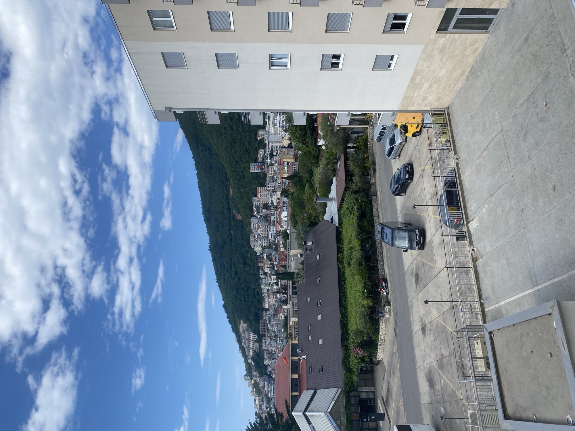 This is an elevated, oblique view of a street and the edge of a building in Budva, Montenegro. The image shows a parking area with several parked vehicles, including a yellow van and a few dark-colored cars. A concrete walkway bordered by a metal railing runs along the parking area. Adjacent to the parking lot, there is a steep hillside densely covered with trees and interspersed with residential buildings. The sky above is blue with scattered white clouds. The architecture of the buildings is contemporary, featuring light-colored facades with a uniform pattern of windows. The overall scene suggests an urban setting with a mix of residential and commercial areas, situated close to natural terrain. There are no visible people in the frame.