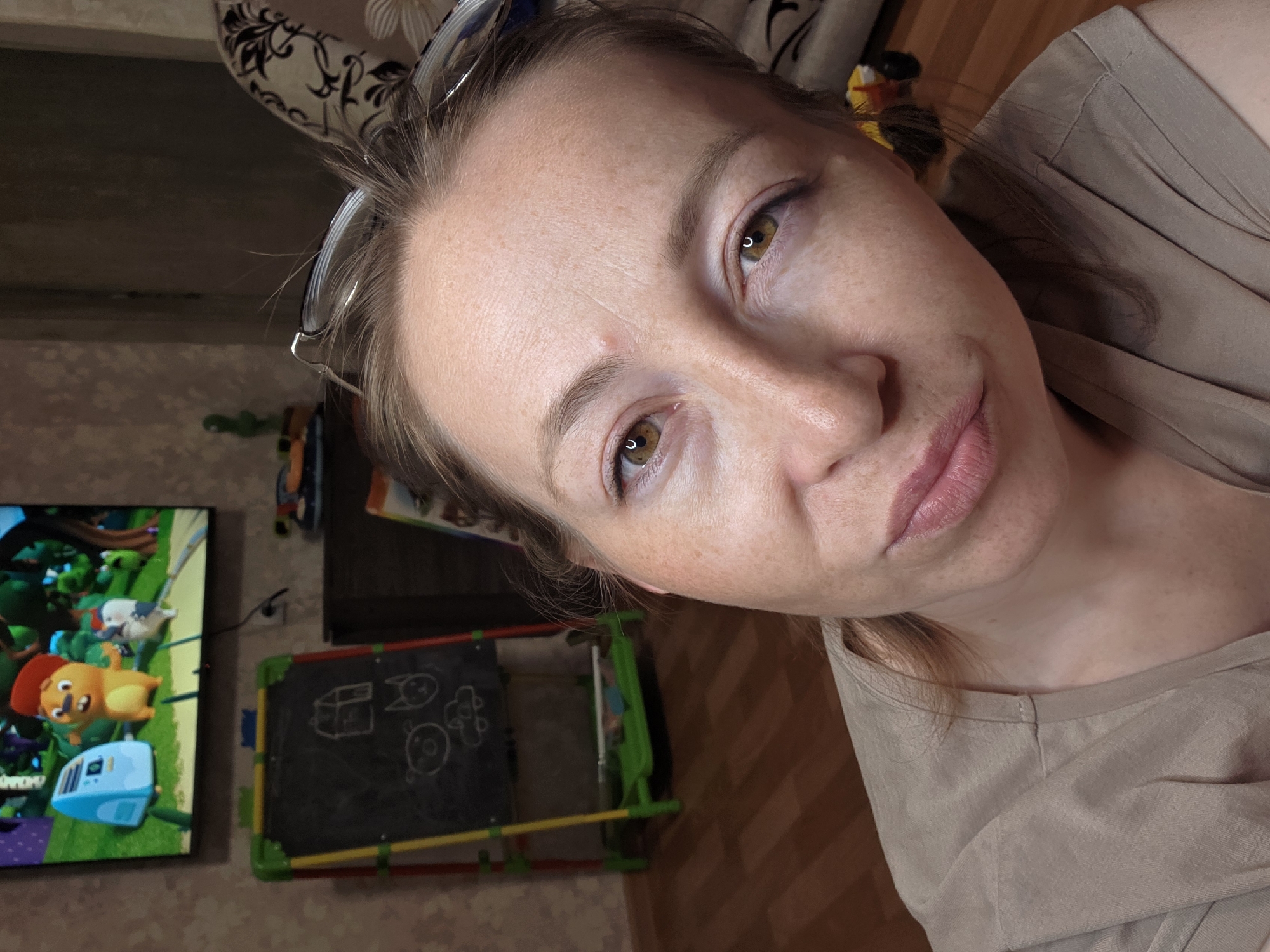 A woman with fair skin, freckles, and brown eyes is pictured in a close-up selfie. She has light brown hair pulled back and is wearing glasses on her head. She is wearing a beige top.
In the background, a television is displaying a cartoon with a yellow animal character. To the right of the television is a child's chalkboard with colorful frames and some basic drawings. The background also includes a patterned cushion and wooden flooring.
The image was captured indoors, likely in a home. The location is presumed to be Chaikovsky, Russia.