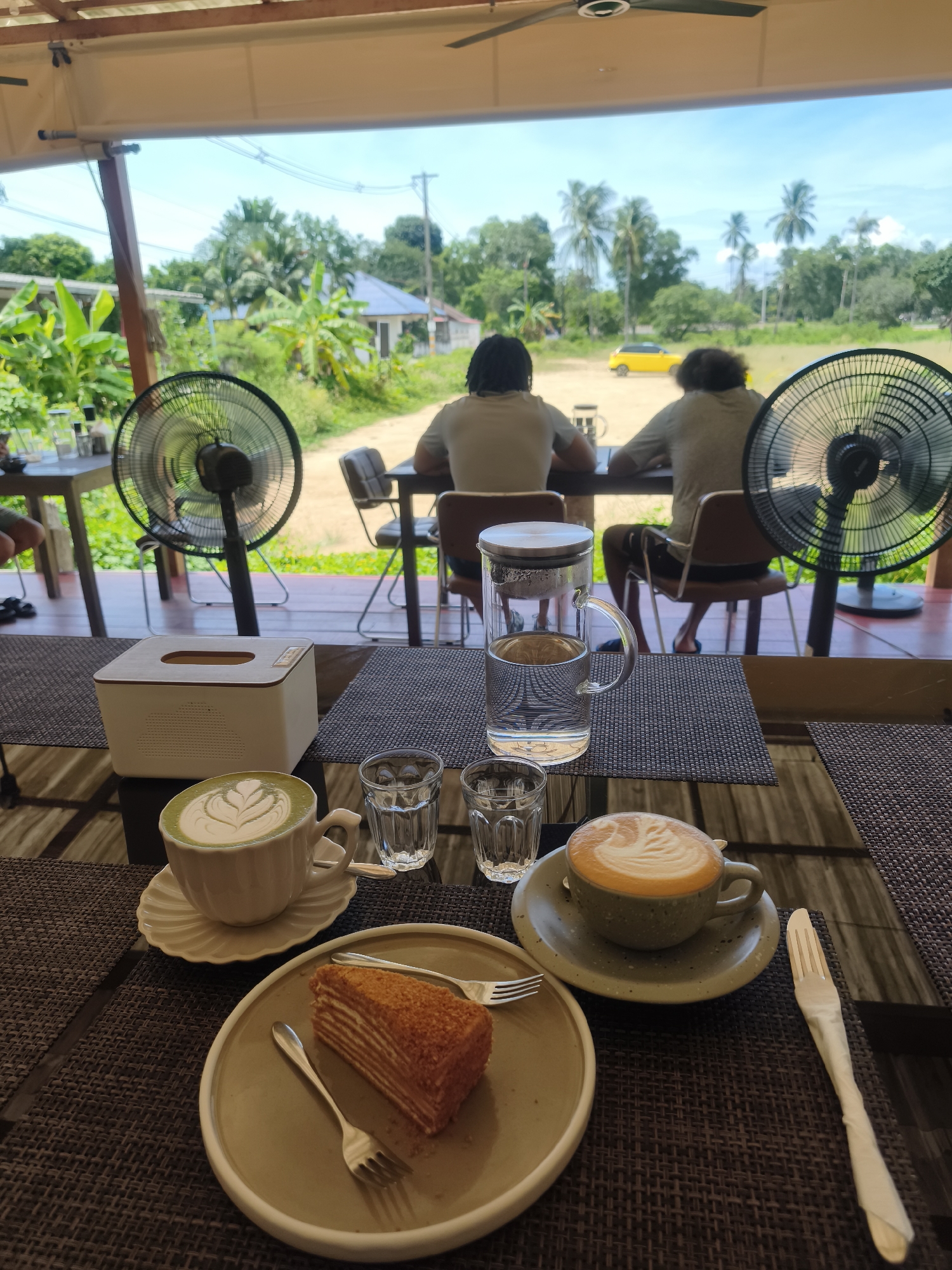 The image depicts a table setting at an outdoor cafe in Baan Mae Nam, Thailand. In the foreground, a table is laid with a brown woven placemat. On it are two cups of coffee, one with green foam art and another with traditional latte art, a slice of layered cake on a plate with two forks, a tissue box, and two small glasses of water. Behind the table, two people are seated at another table, facing away from the camera, looking out at a tropical landscape with palm trees, bananas, and buildings under a bright blue sky. Two black oscillating fans are positioned on either side of the scene, suggesting a warm climate. A yellow car is visible in the distance. The overall scene conveys a relaxed, tropical dining experience.