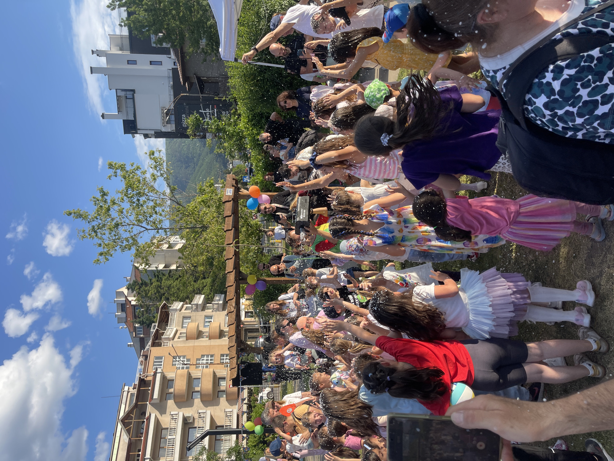 A crowd of people, mainly children, are gathered outdoors under a sunny sky, surrounded by residential buildings and trees. White confetti-like material is being thrown into the air and onto the crowd. Many people are reaching up with their hands toward the falling material. Buildings of different styles and heights surround the event, with trees visible in the background. A wooden structure decorated with balloons stands nearby. The scene likely takes place in a residential area, possibly a park or open space within the city of Sofia, Bulgaria, given the architectural style of the buildings.