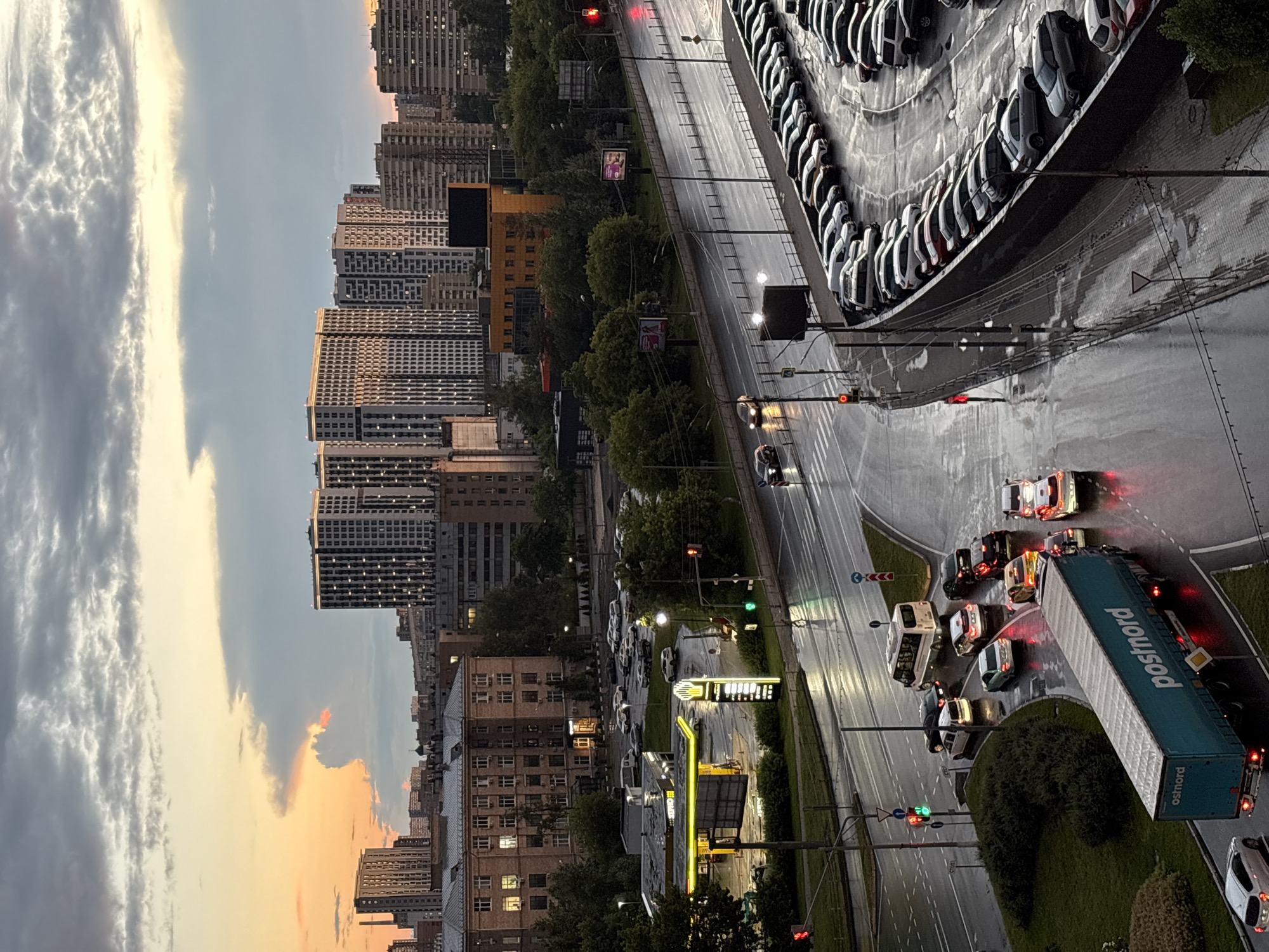 The image depicts a city scene in Mytishchi, Russia, during dusk or dawn, with a dramatic sky featuring orange and gray clouds. A multi-lane road, wet from rain, is visible with several cars and a white bus in transit. The road curves to the right, leading to a large, multi-level parking lot filled with numerous cars. On the left side of the road, a gas station with illuminated pumps and signage is present, along with a group of parked cars. Further in the background, several modern high-rise buildings dominate the skyline. A large teal truck with "postnord" branding is parked on the lower left side of the road, its brake lights illuminated. The overall atmosphere suggests an urban environment in wet weather conditions.