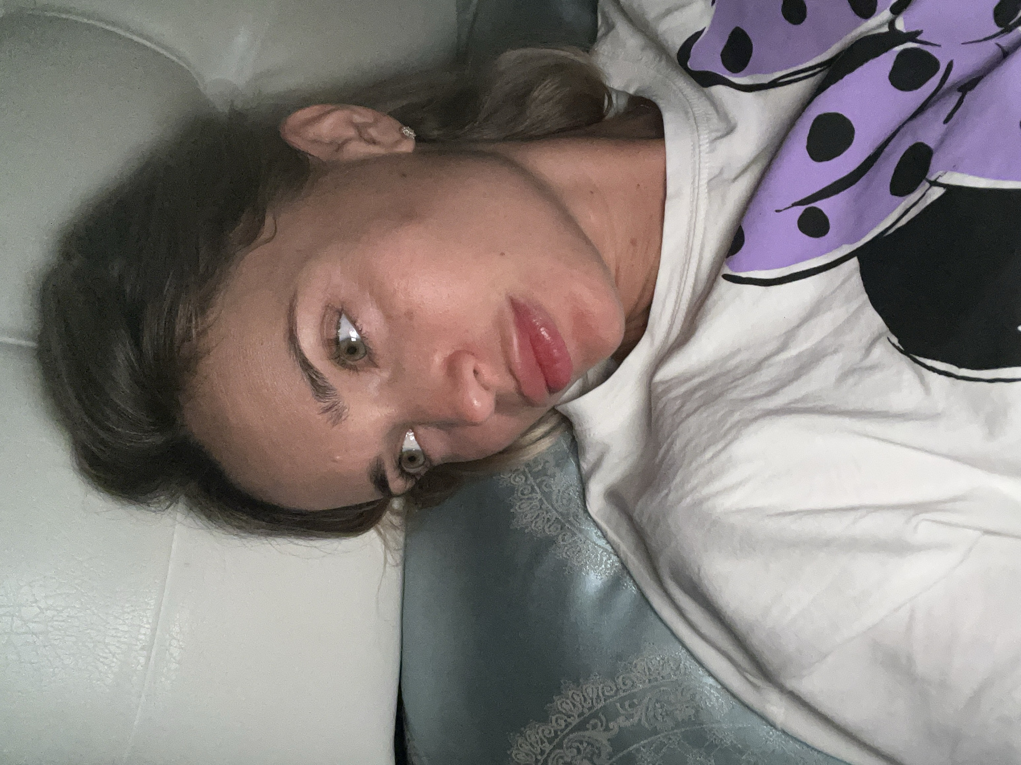 A woman is lying on a bed, with her head on a pillow. She is looking to the side with a pensive expression. She is wearing a white t-shirt with a purple Minnie Mouse bow on it. The room is dimly lit, and the focus is on the woman's face. There is no indication of the location being in the city of Fontanka, Ukraine.