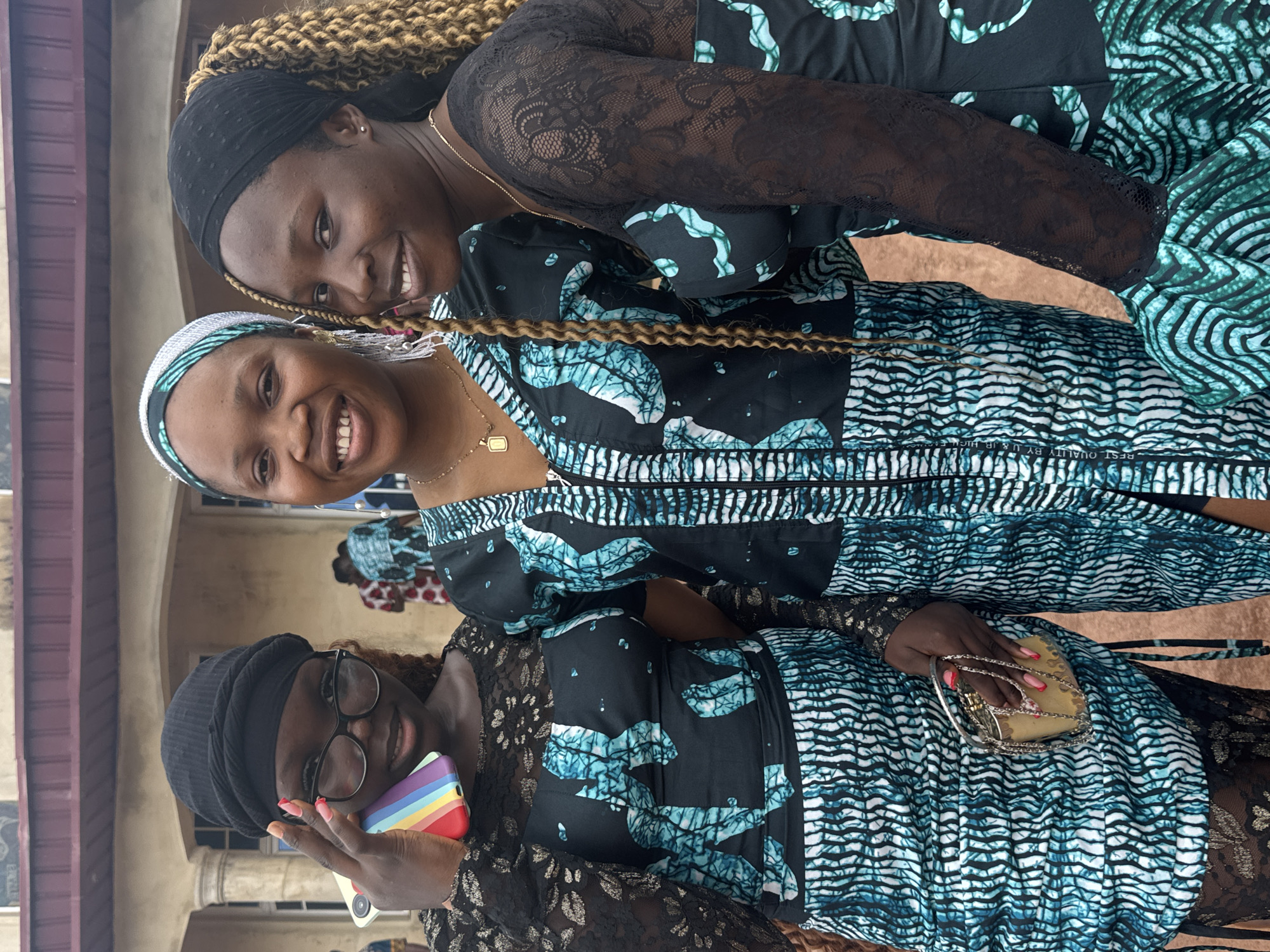 Three women are posing for a photo. They are standing close together, smiling at the camera. Each woman is wearing a similar black and turquoise patterned dress. The woman at the bottom of the frame is holding a rainbow-colored phone case and a small clutch. The woman at the top of the frame has blonde braids and is wearing a black head covering. The woman in the middle has a white and turquoise head covering. The background is slightly out of focus, but appears to be a building in Eziudo, Nigeria.