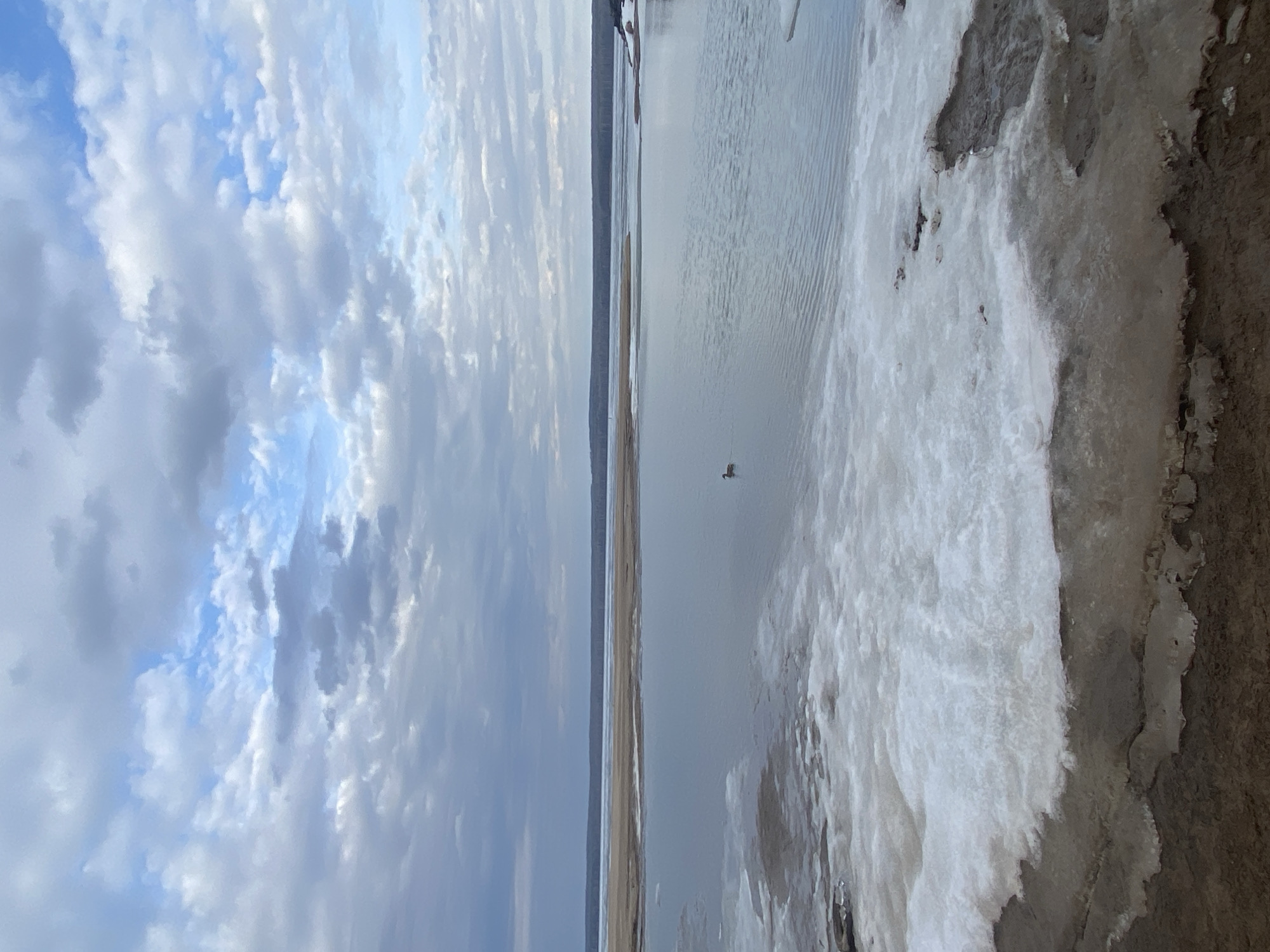 The image displays a wide, serene landscape, appearing to be a river or large body of water, under a partially cloudy sky. The sky, occupying the upper portion of the frame, features a mix of white and grey clouds with distinct patches of blue.
Below the sky, the main body of water stretches across the middle of the image, showing subtle ripples on its surface. A small, dark bird is visible floating on the water, positioned slightly to the right of the center. In the distant background, a thin, dark line extends across the horizon, suggesting a far-off landmass or forest.
The right side of the foreground reveals a shoreline or bank largely covered by ice and snow, indicating cold temperatures, possibly late winter or early spring conditions. The ice appears textured and irregular, contrasting with the darker, exposed ground underneath.
The lighting suggests it is daytime. There are no people visible, nor any discernable text. The scene evokes a sense of tranquility and the vastness of nature.