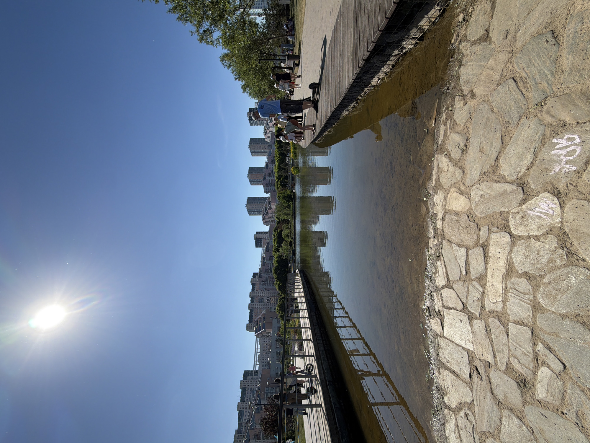 The image depicts a park scene in Khimki, Russia, on a bright, sunny day with a clear blue sky. A strong sun, exhibiting lens flare and streaks, is positioned in the upper left quadrant of the sky.
In the immediate foreground, a path composed of irregularly shaped light-colored flagstones or natural stone tiles is visible, meeting the edge of a shallow, murky body of water. Light-colored graffiti, resembling "OPA," is etched onto one stone in the lower left.
The midground features a calm body of water, reflecting the distant buildings and the wooden boardwalks that line its edges. The water near the shore is visibly shallow and somewhat turbid.
Along the water's edge, two distinct wooden boardwalks are present. On the left, a curved boardwalk with a silver metal railing hosts several individuals walking; one person appears to be pushing a stroller. On the right, a straight section of a wooden boardwalk is occupied by a man in a blue t-shirt and shorts standing beside a dark electric scooter. Adjacent to him, a woman in a white hat leans over the railing, observing the water. Further along this right boardwalk, more people are walking or standing.
The background reveals a series of modern, multi-story buildings with distinct architectural features, including tiered sections, situated across the water and behind a line of green trees and foliage. Reflections of these buildings are discernible in the water. Additional individuals are scattered throughout the park area, walking on paved paths that run parallel to the boardwalks. The overall scene suggests a recreational park environment.