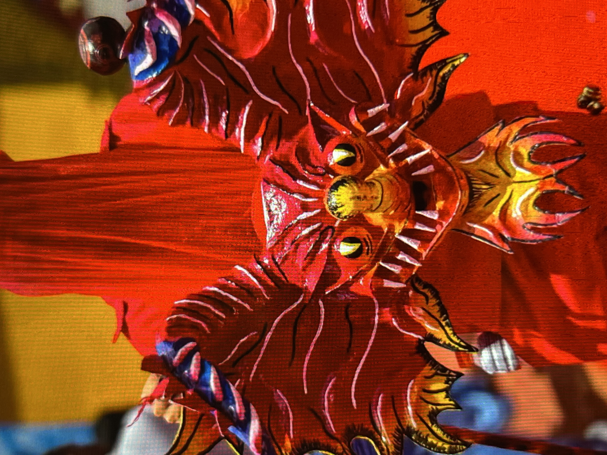 The image shows a close-up of a red, dragon-like mask with yellow eyes and sharp teeth. The mask is ornate, with detailed white and blue accents on its wings. It appears to be part of a costume, as a red fabric is visible behind it. The background is a blurred mixture of yellow and red, suggesting an outdoor event or parade. There are hints of other people in the background, one of whom is wearing a light-colored shirt. The scene is likely from a cultural festival or celebration in Tinaquillo, Venezuela.