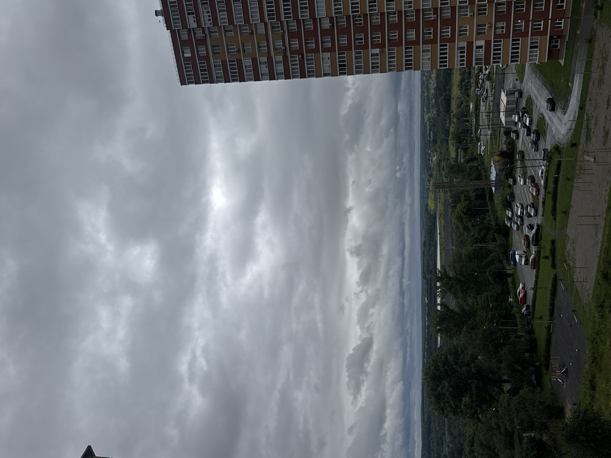 A high-angle, wide shot captures an overcast day in a Russian city, likely Tula, with a modern apartment building dominating the right side of the frame. The building, a multi-story structure with alternating bands of red and yellow brickwork, features numerous windows, air conditioning units, and a visible chimney stack on its roof.
In the foreground, a children's playground with swings and a slide is visible on a patch of gravel and grass. Beyond the playground, a parking lot filled with various cars, including sedans and SUVs, is wet, suggesting recent rain. A small, utilitarian building with a white roof is situated in the parking lot. Streetlights are visible throughout the parking area.
The middle ground is characterized by a dense expanse of green trees, forming a lush treeline that stretches across the landscape. Further in the distance, a body of water, possibly a river or lake, is visible, reflecting the cloudy sky. A tall, metal structure resembling a tower or antenna stands near the water. The horizon is marked by rolling hills covered in trees and scattered buildings.
The sky is filled with heavy, grey clouds, obscuring any clear view of the sun. The overall atmosphere of the image is somber and muted due to the cloudy weather.
