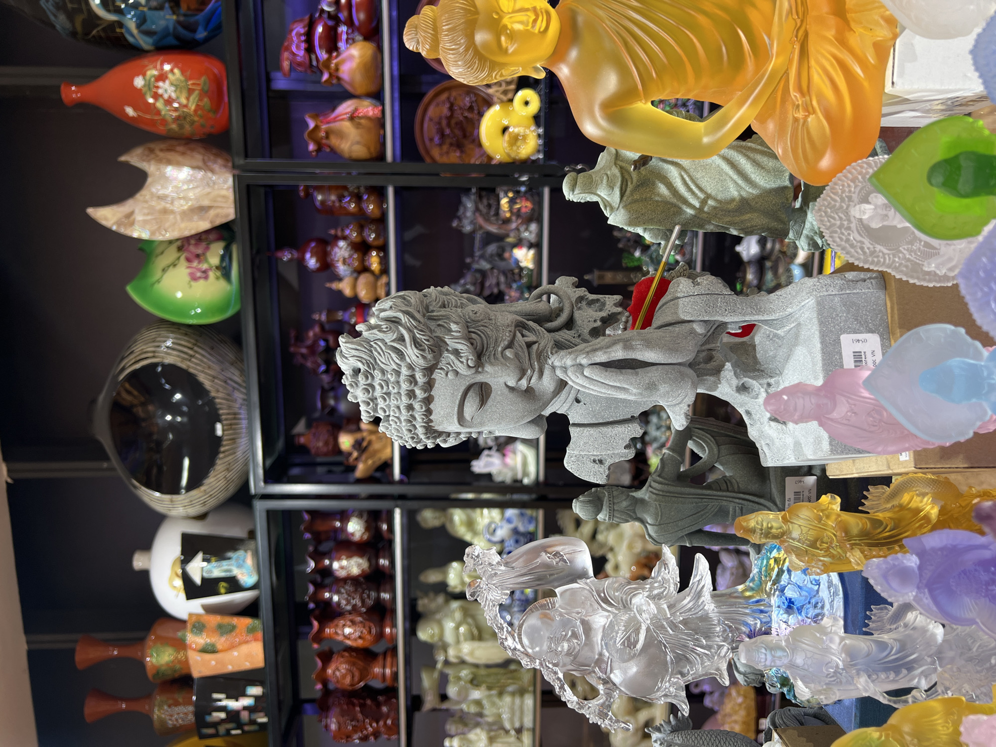 The image shows a display of various decorative objects and statues in a shop or market, possibly located in Nha Trang Ward, Vietnam based on the product label. The foreground features several translucent statues in different colors, including clear, yellow, green, pink, and purple. A stone-like statue of a Buddha-like figure with intricate hair is prominently displayed in the center. The background consists of shelves filled with vases, pottery, and other decorative items, creating a crowded and diverse display. The lighting suggests an indoor setting, and the overall impression is that of a retail environment selling souvenirs and decorative arts.