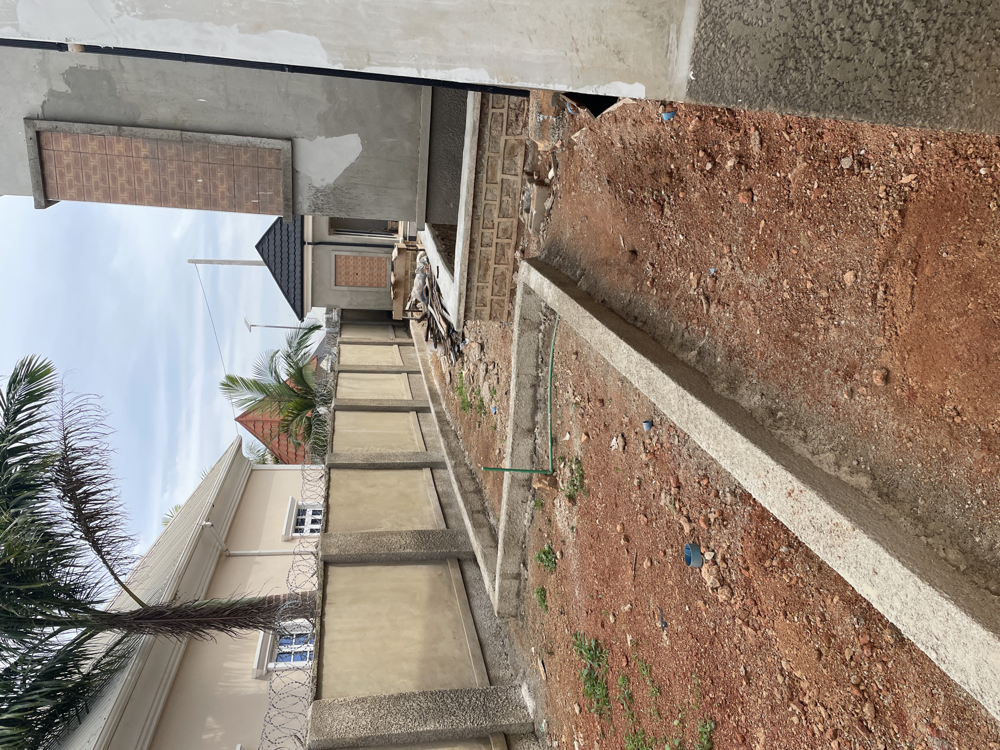 In Bar Gada, Nigeria, a construction site is depicted, characterized by red soil and incomplete building structures. A beige-colored wall, topped with barbed wire, runs horizontally across the mid-ground, separating the foreground from the background. Behind the wall, a pale yellow building with white trim is visible, featuring multiple windows. To the right of this building, a darker structure with a black, pitched roof is under construction. In the foreground, a low concrete barrier outlines a section of the site, and more red soil is present, interspersed with small green plants and scattered debris. Another, partially constructed building is visible on the far right, with unfinished grey concrete walls. The sky above is overcast.