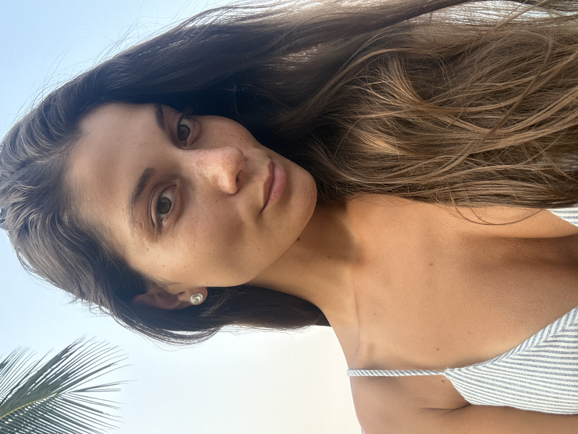 A young woman with long, brown hair and fair skin is depicted in a close-up selfie. Her face is angled slightly, and she looks directly at the camera. She has brown eyes, freckles on her nose, and is wearing a small, round earring. She is wearing a striped garment with thin straps. A section of a palm frond appears in the lower left corner of the frame, suggesting a tropical or coastal location. The background is a clear, light-blue sky. The picture provides no explicit information about Đà Nẵng, Vietnam.