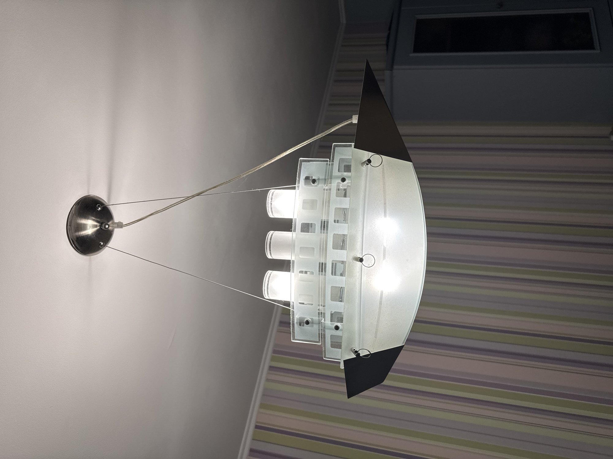 An upside-down, frosted glass light fixture shaped like a cruise ship hangs from a ceiling in a room. The light fixture has multiple levels, with cylindrical elements that appear to be smokestacks at the top. The ship's hull is a wide, curved frosted glass, and it has a dark, pointed bow. Two small rings are attached to the sides of the hull. The ceiling is white, and a metal canopy at the top anchors the wires suspending the light. The background features a wall with vertical striped wallpaper in muted shades of green, pink, and purple, and to the right, a portion of a tall, dark blue cabinet is visible. The lighting in the room is dim, with the ship fixture being the primary light source. The overall scene suggests an interior space, possibly a residential room.