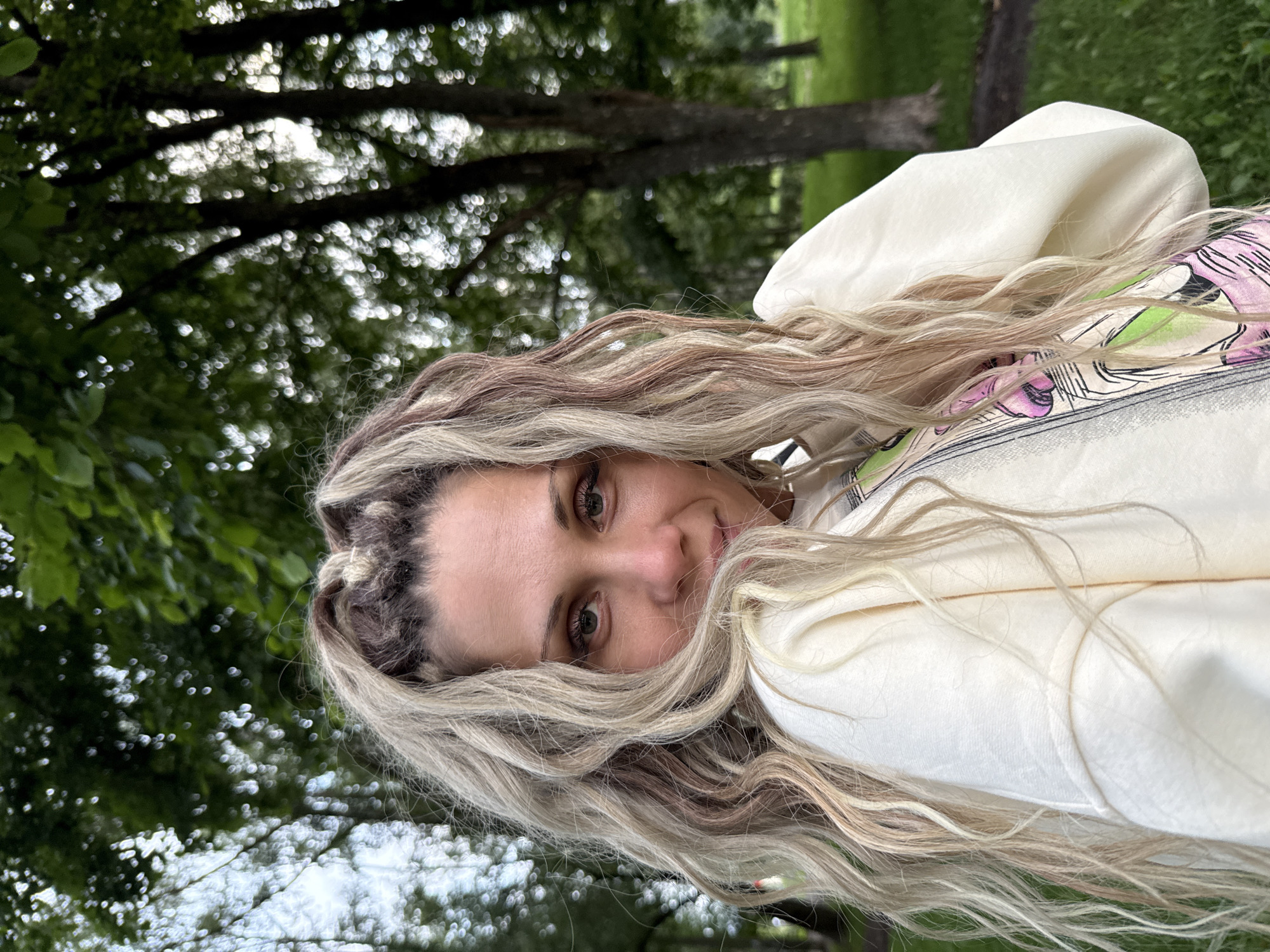 A medium shot captures a woman with long, blonde, and brown braided hair styled in dreadlocks, her face turned slightly towards the viewer, in what appears to be a park. She is wearing a cream-colored sweatshirt with a graphic design featuring pink and green elements. The background consists of a blurred forest with green foliage and tall trees, indicating an outdoor, natural setting. The lighting is soft, suggesting an overcast day or late afternoon. The woman's eyes are looking directly at the camera with a gentle expression. The scene is devoid of any overt actions or interactions, focusing solely on the woman's portrait. The location is specified as Rusanovo, Russia.