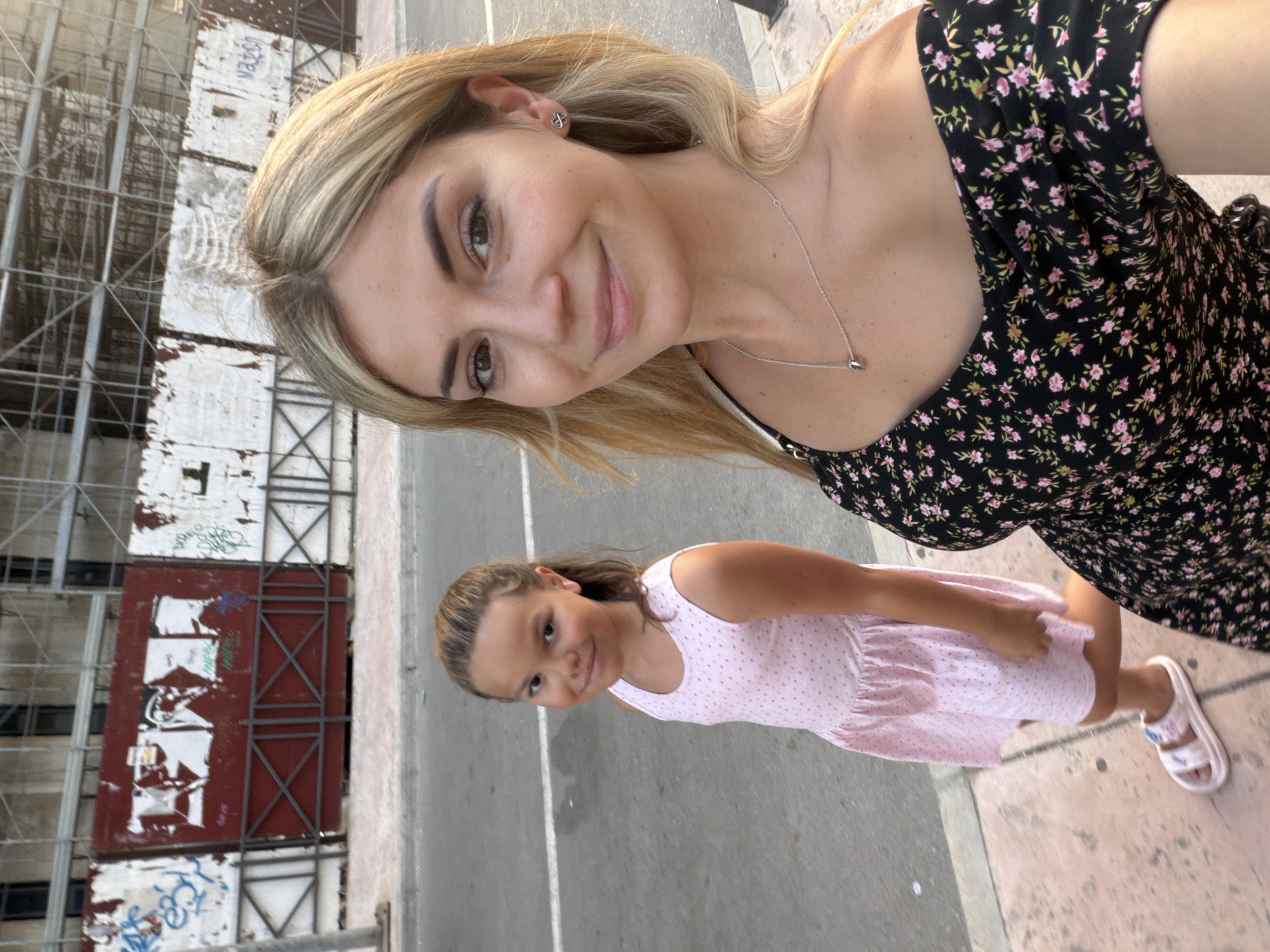In Budva, Montenegro, a woman and a young girl are walking on a sidewalk next to a street. The woman is taking a selfie, with the girl slightly behind her. The woman has blonde hair and is wearing a black floral-print off-the-shoulder dress and a silver necklace with a teardrop pendant. The girl has brown hair pulled back in a ponytail and is wearing a pink polka-dot dress and white sandals. Behind them, a building under construction is visible, with scaffolding and white construction barriers covered in graffiti. The street appears to be a main road, with a white line marking the lane. The lighting suggests it is daytime, possibly late afternoon.
