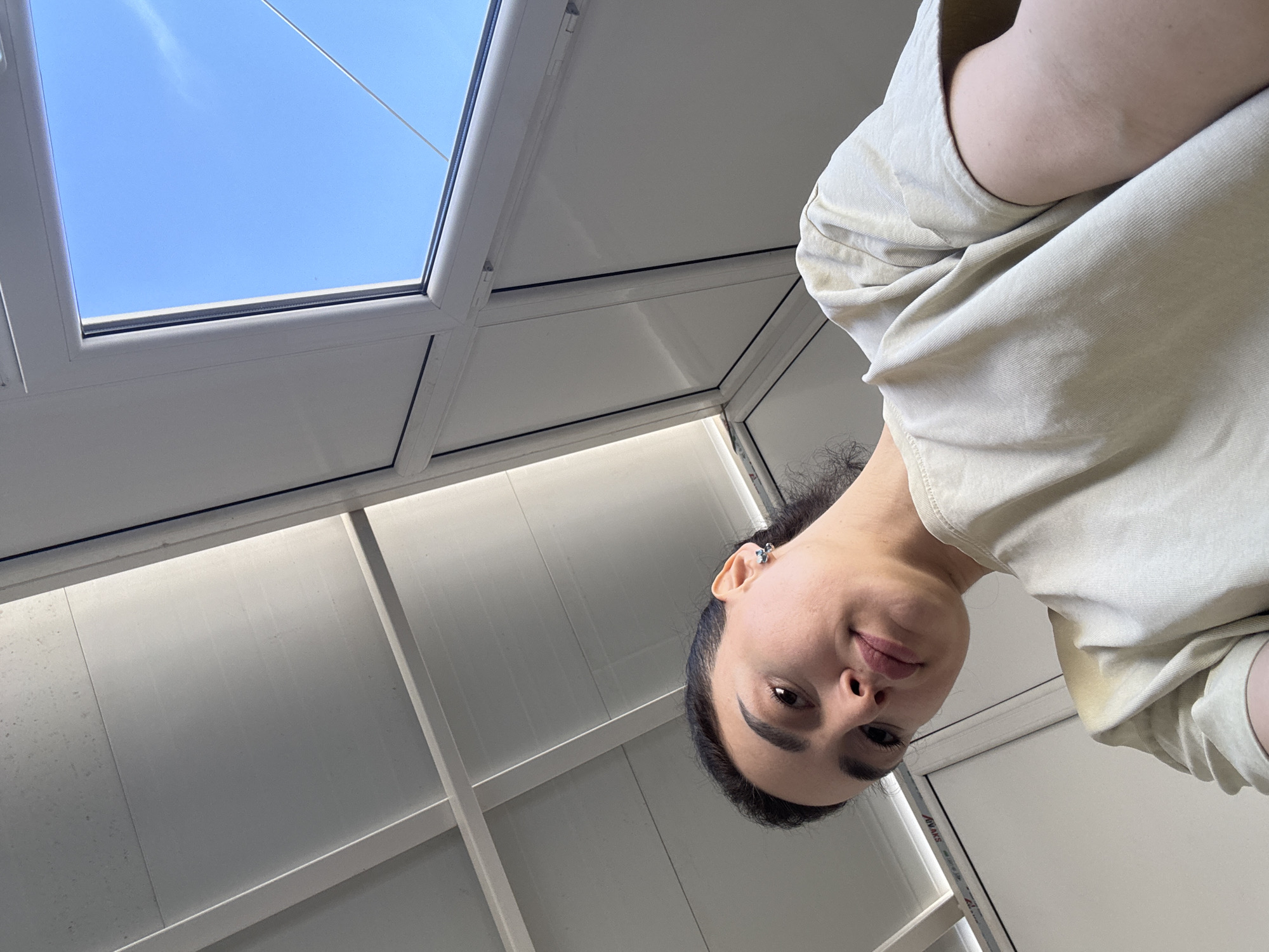 The image shows a person looking down at the camera, with a view of a blue sky through a skylight in the background. The person has dark hair pulled back and is wearing a light-colored top. The skylight is framed by white panels, and the overall setting appears to be indoors. The location, Tbilisi, Georgia, is not discernible from the image.