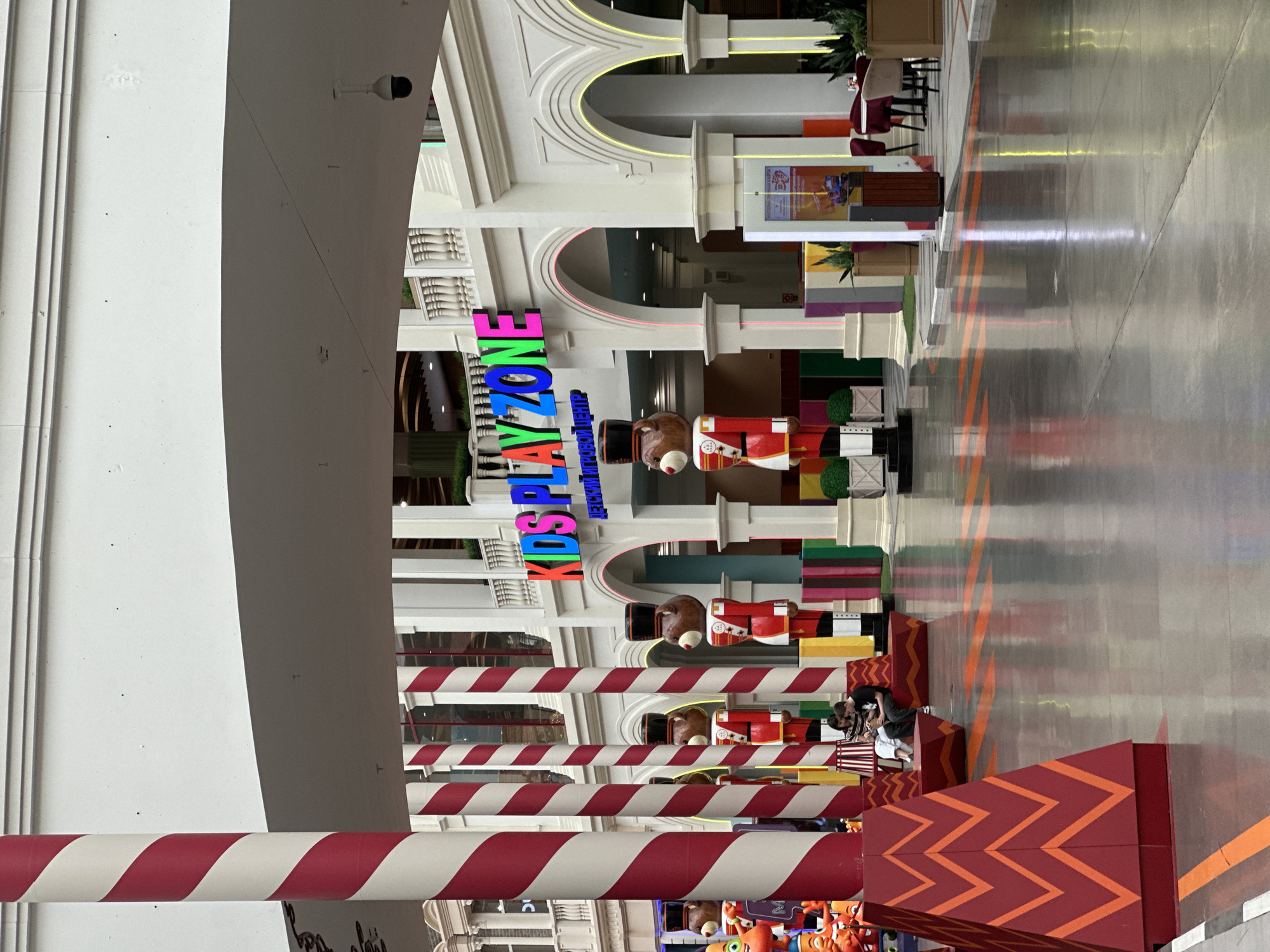This image depicts an indoor scene within a large commercial complex, likely a shopping mall or entertainment center, in Moscow, Russia. The central focus is a vibrant, multicolored sign that reads "KIDS PLAYZONE" with smaller text "Grand Kanyon" underneath, indicating a children's play area.
In front of the "KIDS PLAYZONE" sign, several large decorative figures resembling nutcrackers with teddy bear heads and red, gold, and white soldier uniforms are arranged, adding a whimsical and possibly festive atmosphere. The foreground is marked by multiple red and white candy-cane striped poles or barriers and large red rectangular blocks adorned with orange zigzag patterns, which appear to be part of the play area's entrance or boundary. A few people are visible in the lower right corner, near these striped barriers, appearing to be adults or older children.
The architectural style is grand, featuring multiple levels with white columns, classical arches, and balustrades. Some arches are highlighted with decorative yellow and pink neon lighting. On an upper level, tables and chairs can be seen, suggesting nearby dining or lounge areas. The floor is a polished surface with subtle orange linear patterns. A security camera is mounted on the ceiling. The space is brightly lit, indicative of an indoor daytime or evening setting, designed to be inviting and engaging for visitors, particularly families with children.