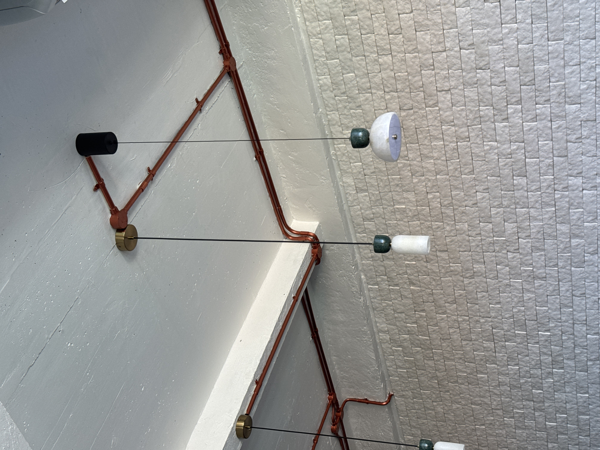 The image provides an upward view of an interior space, likely a ceiling or upper wall, in Kuta Selatan, Indonesia, showcasing a modern or industrial design aesthetic. The setting features a textured, off-white wall surface on the left, juxtaposed against a section of light grey, rectangular, brick-like tiles on the right. Exposed reddish-brown electrical conduits are prominent, running across the white wall surface in angular patterns, contributing to the industrial feel.
Several minimalist light fixtures or decorative elements are suspended by thin black wires. Each suspension point on the white wall includes small, round, brass-colored fittings, from which the wires extend. A black cylindrical element is also visible as part of the upper-left suspension. Each suspended element consists of a dark green, oblong piece. The topmost fixture has this green piece connected to a white, semi-spherical component. The two lower fixtures feature the dark green oblong piece connected to a white cylindrical component.
No people or specific activities are depicted in the image. The focus is solely on the architectural and lighting details. There is no visible text.