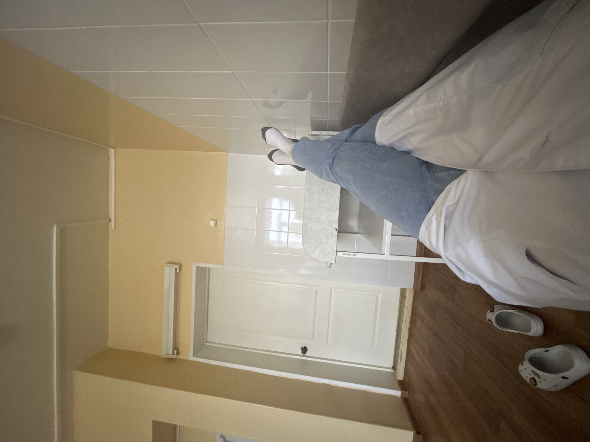 The image captures an indoor scene, likely a break room or office within a medical or institutional setting, given the presence of a lab coat and the overall utilitarian decor. The perspective is a first-person view, looking down towards one's feet and across the room.
A person is seen reclining on a dark grey couch or bench, with their legs extended. They are wearing light blue denim jeans and white socks with dark grey or black toes and heels. A white lab coat or similar professional garment is draped over their lap and lower body. On the wooden floor near the bottom right, a pair of white clogs, possibly Crocs, with some subtle markings, are placed.
The room features a diverse floor covering, transitioning from light brown wooden planks in the foreground to white square tiles further back, beneath the person's feet. The walls are two-toned; some sections are painted a light beige or pale yellow, while others are covered in white square tiles, particularly behind where the person's feet are positioned. A white panel door with a silver handle is visible in the background, set into a wall. Above the door, a rectangular fluorescent light fixture is mounted. A small white cabinet or shelf unit is also present in the background. The ceiling features white acoustic tiles.
On the white cabinet, a label with the text "21013600019" is visible. The scene suggests a moment of rest or downtime for the individual in a functional environment in Фоки, Russia.