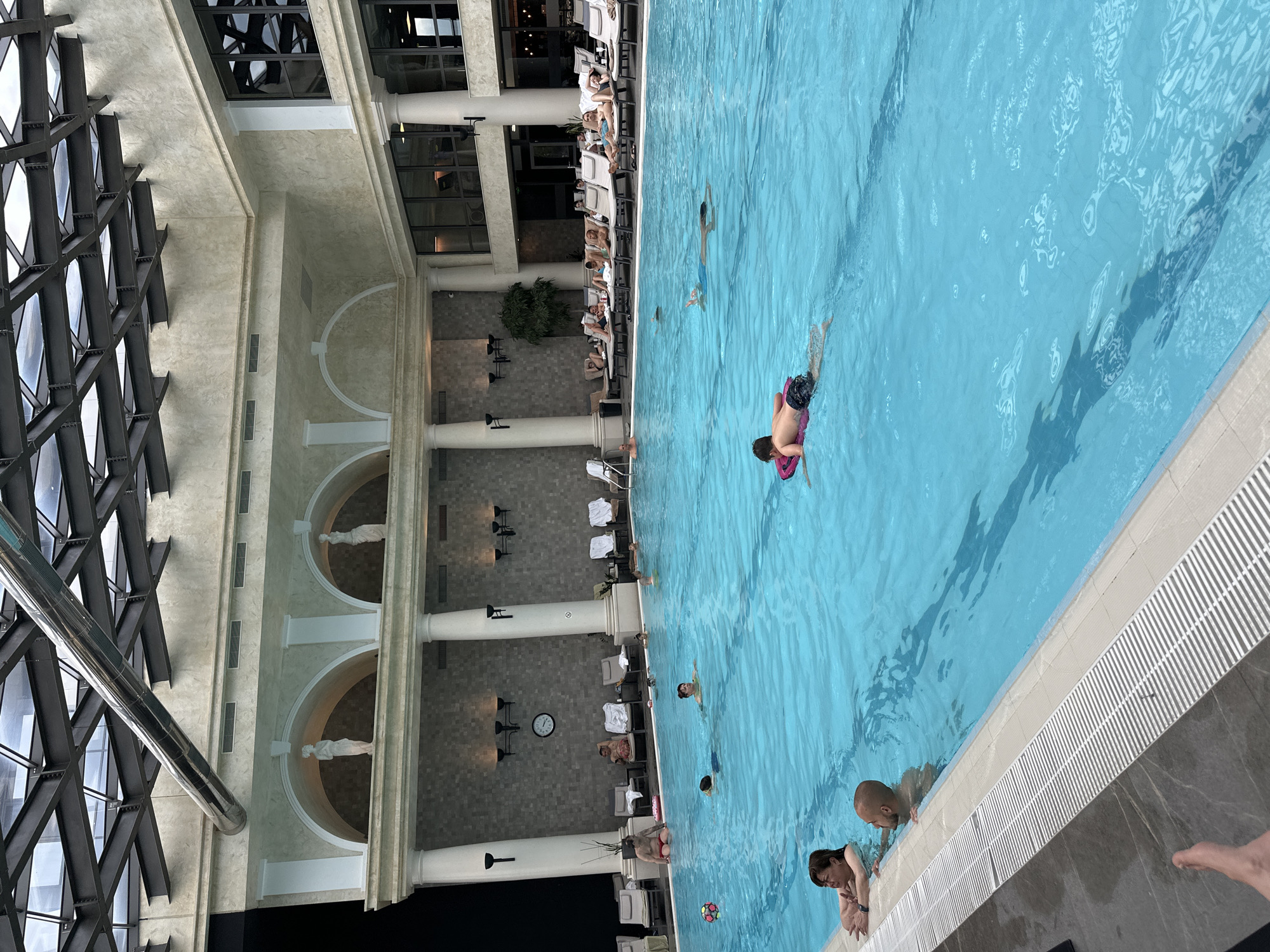 The image shows an indoor swimming pool in Kazanlak, Bulgaria. The pool is rectangular and filled with blue water, with several people swimming and standing near the edge. Some individuals are using inflatable devices. Adjacent to the pool is a tiled area with lounge chairs where people are relaxing. The pool area is indoors, with a high ceiling supported by columns. Statues are positioned in the arches between the columns. The building has large windows and a metallic framework structure.