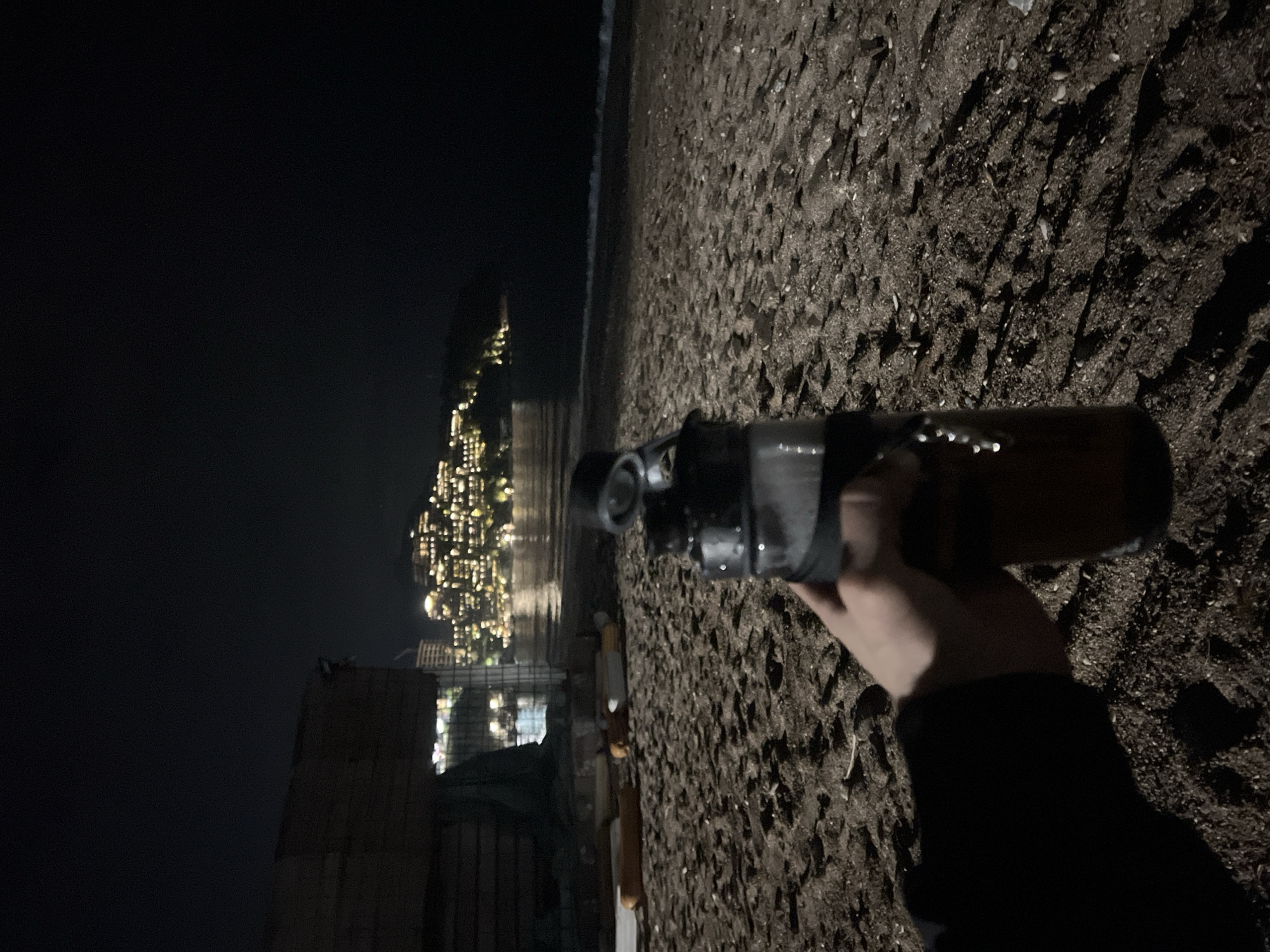 The image shows a hand holding a bottle, possibly modified, at night on a dark beach. In the background, across the sea, is the city of Bečići, Montenegro, illuminated with numerous lights. To the left, there's a partial view of a structure, possibly a building or wall, made of dark materials. The beach surface is rough and textured. The person is wearing dark clothing.