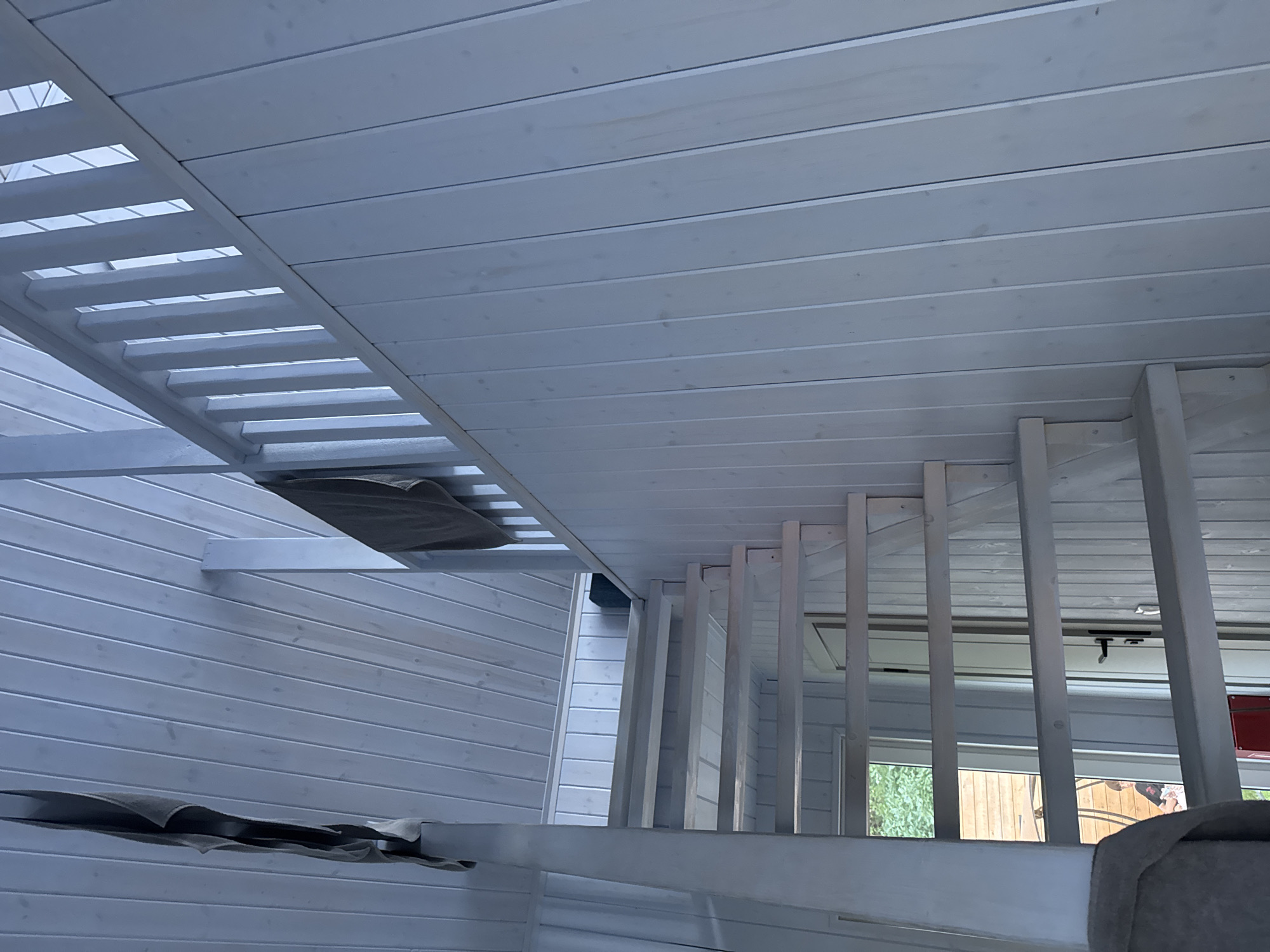An interior shot shows a wooden staircase with white painted planks. The staircase is located in a building with white planked walls and a white planked ceiling. A dark grey towel hangs from the railing of the staircase on the left side, and another dark grey towel hangs from the railing on the right side. The bottom of the staircase leads to a doorway with a wooden door and a black door handle. Through the doorway, a person is visible sitting on a chair. The environment suggests a residential or recreational setting, possibly a cabin or a home with a loft. The overall impression is one of a simple, clean, and unfinished interior space. The location is Doma 1096 Km, Russia.