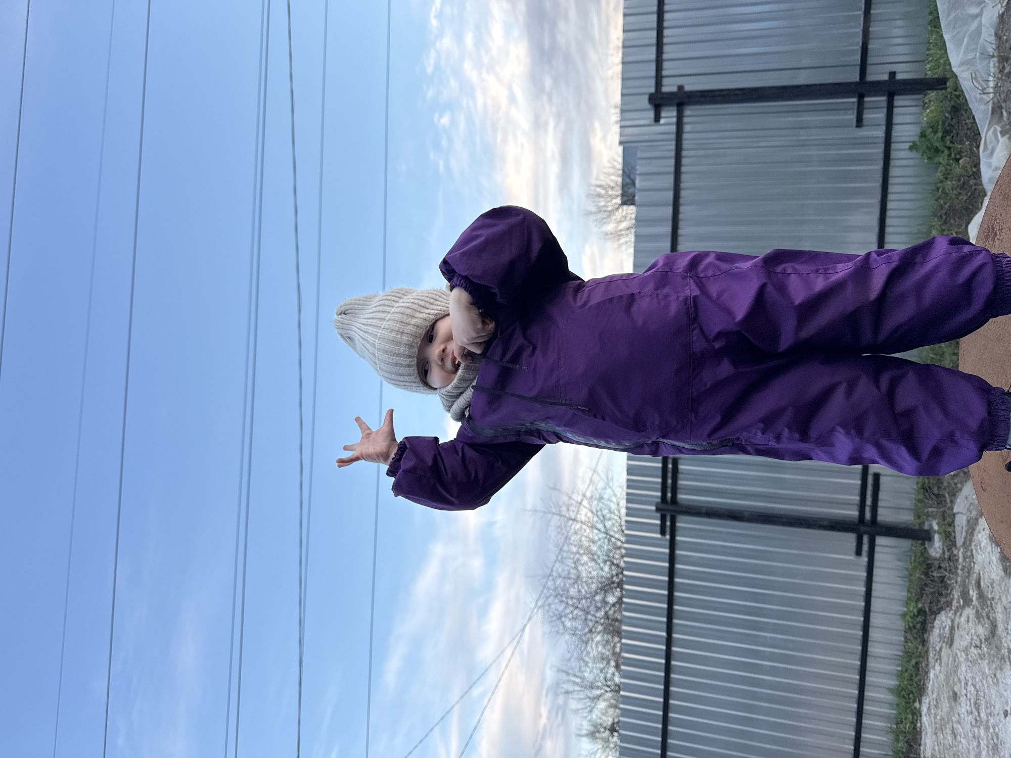 A young child, likely a toddler, stands upright outdoors, looking towards the camera with a subtle expression. The child is warmly dressed in a dark purple hooded full-body snowsuit and a light grey knitted beanie. One hand is raised with fingers splayed open in a playful gesture, while the other is near their face.
The background is divided between an expansive light blue sky with scattered white clouds on the left, featuring several thin power or telephone lines stretching horizontally across the frame. On the right, a corrugated metal fence, supported by black metal posts and horizontal bars, provides a stark backdrop. Some dry grass or weeds are visible at the base of the fence, and bare tree branches can be faintly seen through and above it. The child appears to be standing on a reddish-brown, circular object, possibly a planter edge or another ground-level structure, partially visible in the bottom right corner.
The scene depicts a daytime outdoor moment on what appears to be a cool, partially cloudy day. There is no visible text in the image. The location is Chaikovsky, Russia.