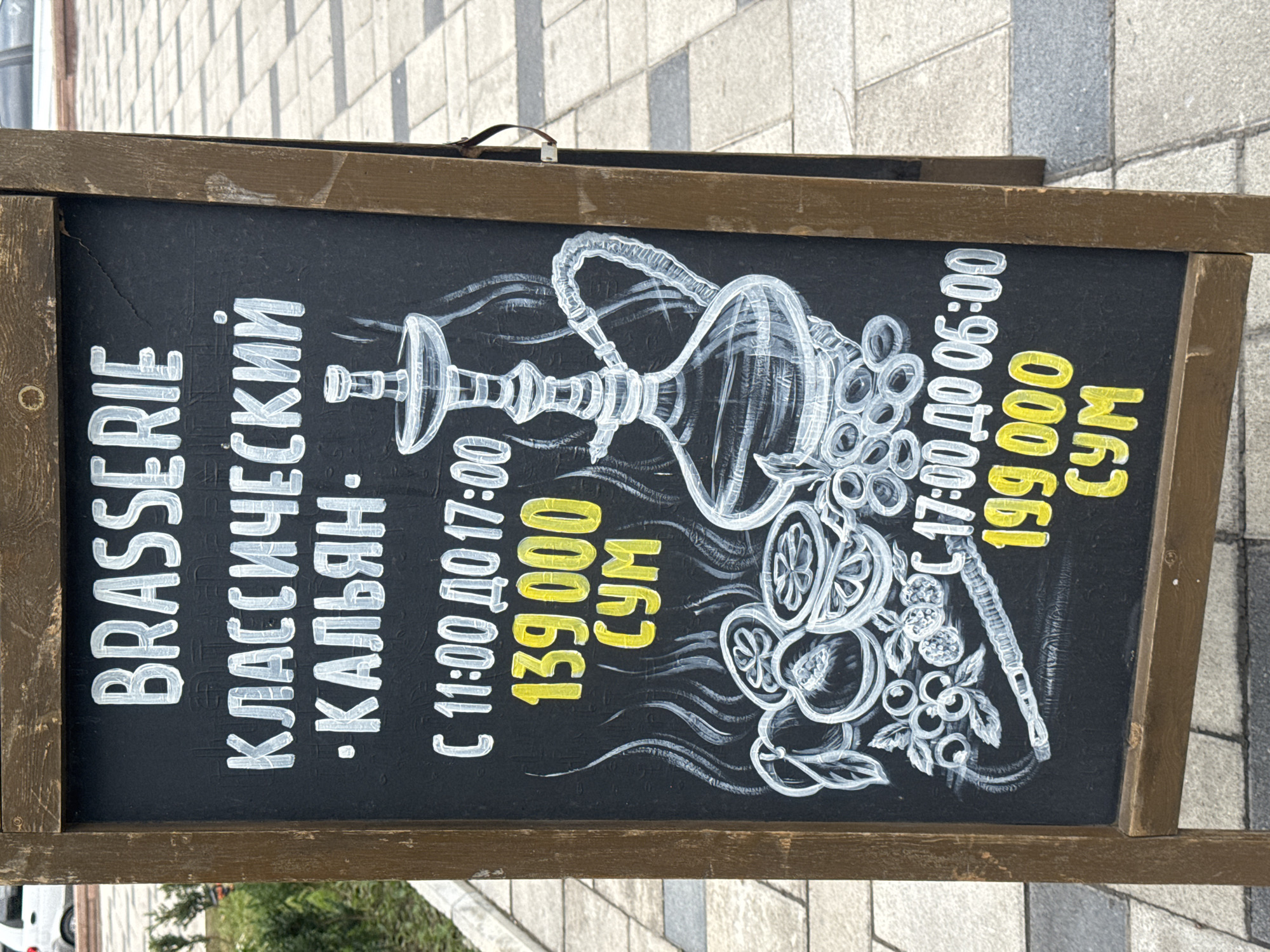 This is a horizontal, eye-level shot of a chalkboard sign advertising shisha (hookah) prices at a brasserie in Tashkent, Uzbekistan. The sign is propped up on a sidewalk with a tiled pattern. A wooden frame surrounds the black chalkboard.
The chalkboard is illustrated with a white drawing of a hookah, complete with a base, hose, and bowl. Various fruits, such as lemons and berries, are also depicted around the hookah. White lettering at the top of the sign reads "BRASSERIE" in English, followed by "КЛАССИЧЕСКИЙ • КАЛЬЯН" in Cyrillic, meaning "CLASSIC • HOOKAH".
Below the illustration, the sign details two price tiers for the hookah service:
* "С 11:00 ДО 17:00" (From 11:00 to 17:00) is priced at "139000 СУМ".
* "С 17:00 ДО 06:00" (From 17:00 to 06:00) is priced at "199 000 СУМ".
The time of day appears to be daytime, likely late morning or early afternoon, given the lighting and the context of the price structure. The weather seems fair, as the sign is displayed outdoors without any apparent protection from the elements. The setting is a public area, possibly an outdoor seating area for the brasserie. No people are visible in the image.