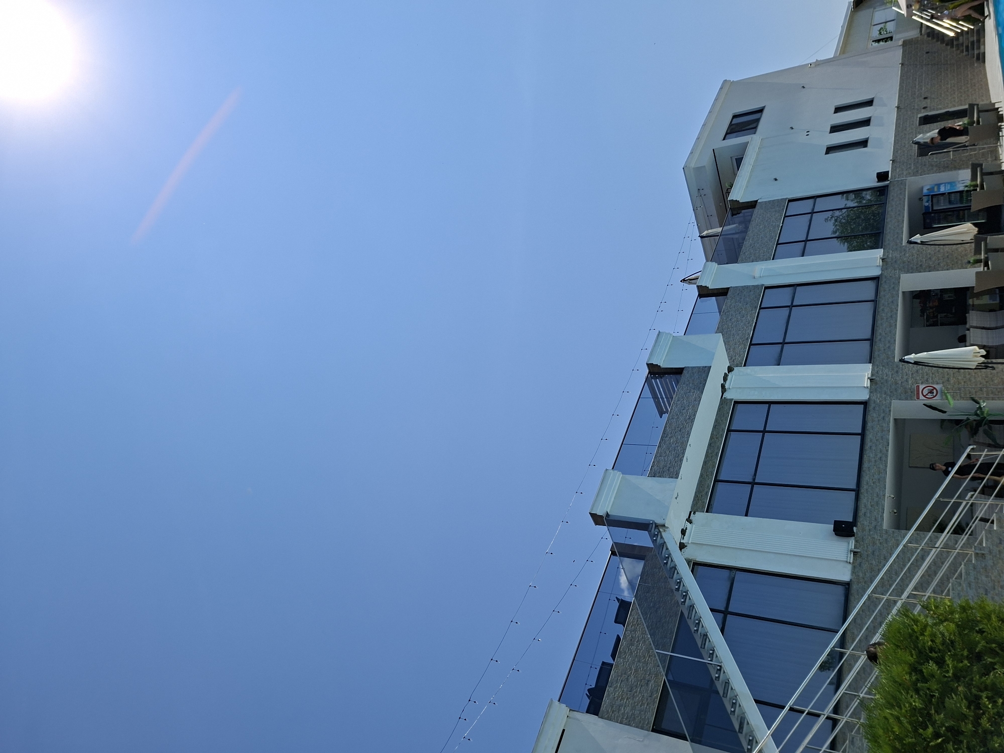 In Costești, Moldova, a modern, multi-story building with large reflective windows and a stone facade is viewed from a low angle against a clear blue sky. A bright sun is visible in the upper right corner of the sky, casting a lens flare across the image. String lights with small bulbs are strung across the upper section of the building. A metal staircase with glass railings ascends along the left side of the building, leading to an upper level with outdoor seating and umbrellas. In the foreground, a portion of a green shrub is visible. Several tables and chairs are set up outside the building, some with white umbrellas providing shade. A person in a black t-shirt and shorts is walking near the entrance to the building, and another person in a white shirt and shorts is seen walking up the outdoor stairs on the right side of the image. The architecture suggests a hotel, restaurant, or resort.