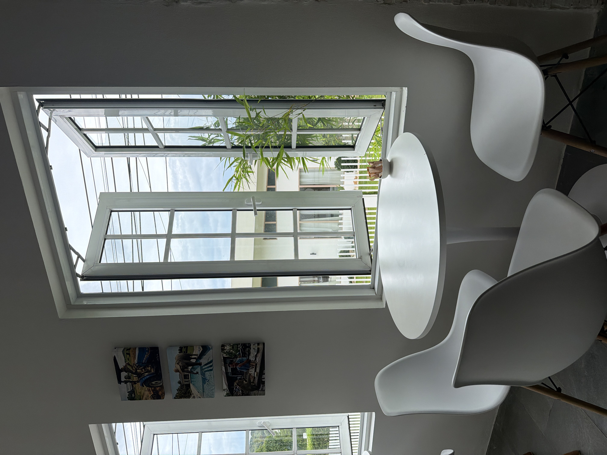 This image depicts an indoor seating area with a round white table and three white chairs. The table has a small white bowl with what appear to be sugar packets on it. Behind the table is a large window with multiple panes, which is open, allowing a view of greenery and a building outside. On the wall to the left of the window, there are three square framed pictures stacked vertically. The first picture shows a person in a blue tractor. The second shows an elephant by a swimming pool. The third picture shows a person playing a board game with a monkey. The room has white walls and a dark grey floor. The overall setting appears to be a cafe or restaurant in Ko Samui District, Thailand, judging by the style of the building visible outside and the presence of tropical vegetation.