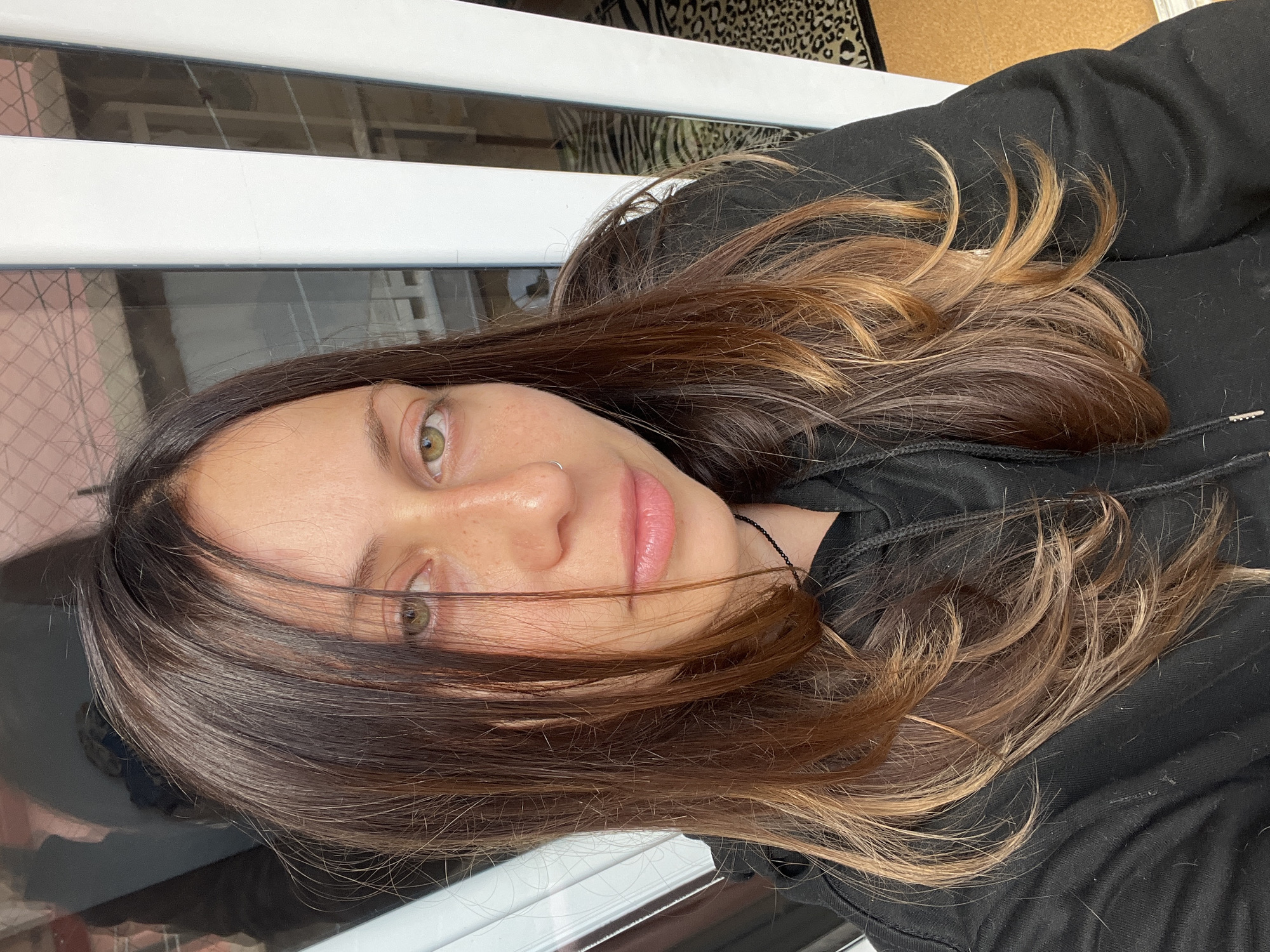 A close-up portrait shows a young woman looking directly at the camera. The image is oriented with her head on the left side, indicating a 90-degree counter-clockwise rotation from a typical upright perspective. She has long, wavy hair with a distinct balayage style, featuring dark brown roots that transition into lighter brown and caramel highlights towards the ends. Several strands of her hair fall casually across her face.
Her eyes are green or hazel, and she has light freckles visible across her nose and cheeks. Her lips are a natural pink color, and she wears a small silver hoop nose ring on her right nostril. She is dressed in a black hoodie or jacket, with a thin black cord visible around her neck. Her expression is relaxed and neutral.
The background behind her features white horizontal slats, which could be window blinds or a balcony railing, reflecting what appear to be urban buildings with grid-like windows. In the upper part of the background (which would be the top right if the image were upright), a leopard print pattern on fabric and a light brown, possibly cork, surface are partially visible. The lighting is bright and natural, suggesting the photo was taken during the daytime. No text is visible in the image. The urban setting is consistent with Barcelona, Spain.