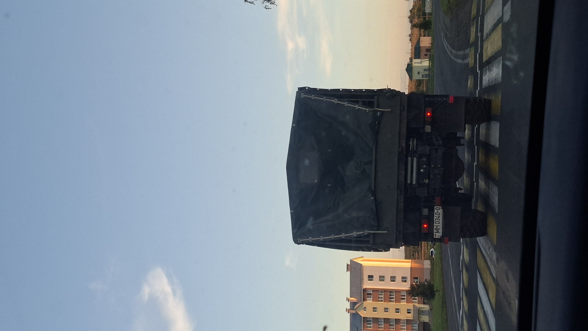 The image, taken in Myadzyel, Belarus, shows the rear view of a military truck crossing a pedestrian crossing. The truck is dark green and covered with a tarp. Its license plate reads "MH 0340-0". Red taillights are illuminated. In the background, there are apartment buildings, a few smaller houses, and a sparse treeline under a clear blue sky with some wispy clouds. The pedestrian crossing has alternating yellow and white stripes. The perspective suggests the photo was taken from inside another vehicle.