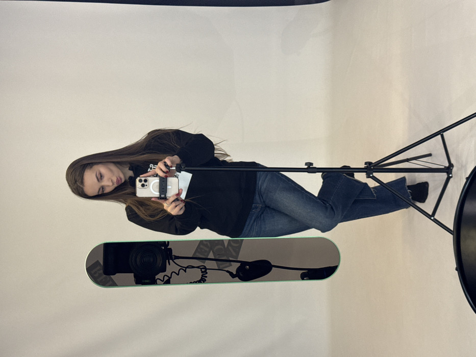 A young woman in a black sweatshirt and blue jeans takes a selfie with a smartphone. The phone has a white ring around the camera and is held in her right hand. She appears to be in a photography studio, with a light stand visible behind her and a snowboard on the floor reflecting studio equipment. The background is plain white, typical of a photo studio setting.