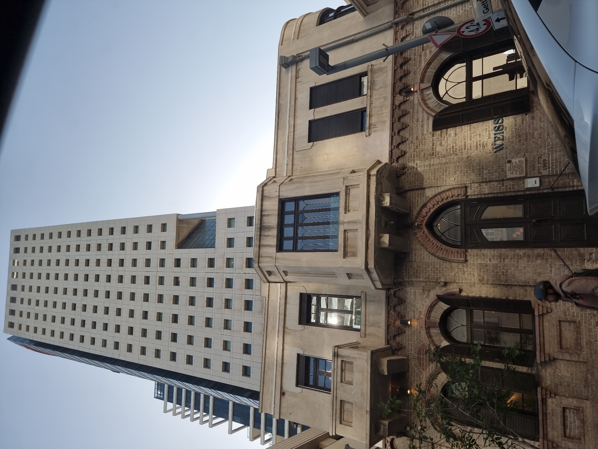 The image presents a street view in Tel Aviv, Israel. On the right, an older building is constructed with brick and beige accents featuring arched windows and the signage "WEISS". The building is attached to a newer structure with a white facade punctuated by numerous square windows. A street sign is visible above the right building. A vehicle appears partially on the right edge of the image, and a person is vaguely visible near the bottom right corner. The sky is light blue.