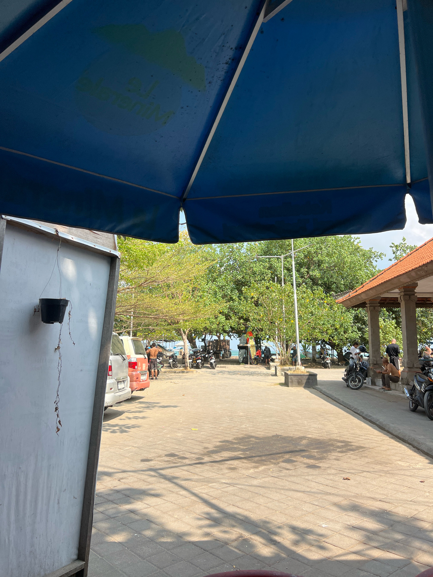 An outdoor paved area in Kuta, Indonesia, with a large blue tarp overhead, partially obscuring the view of a tropical environment. In the foreground, a white structure is visible on the left, with a black hanging planter. Behind it, a silver SUV and a red van are parked, with two men standing between them. The background reveals lush green trees and a glimpse of the ocean. Several motorcycles are parked throughout the scene, with people around them. To the right, a traditional Balinese building with a tiled roof and stone pillars stands. The ground is paved with interlocking concrete blocks, and shadows of trees are cast across it. The overall atmosphere suggests a busy beachfront area.