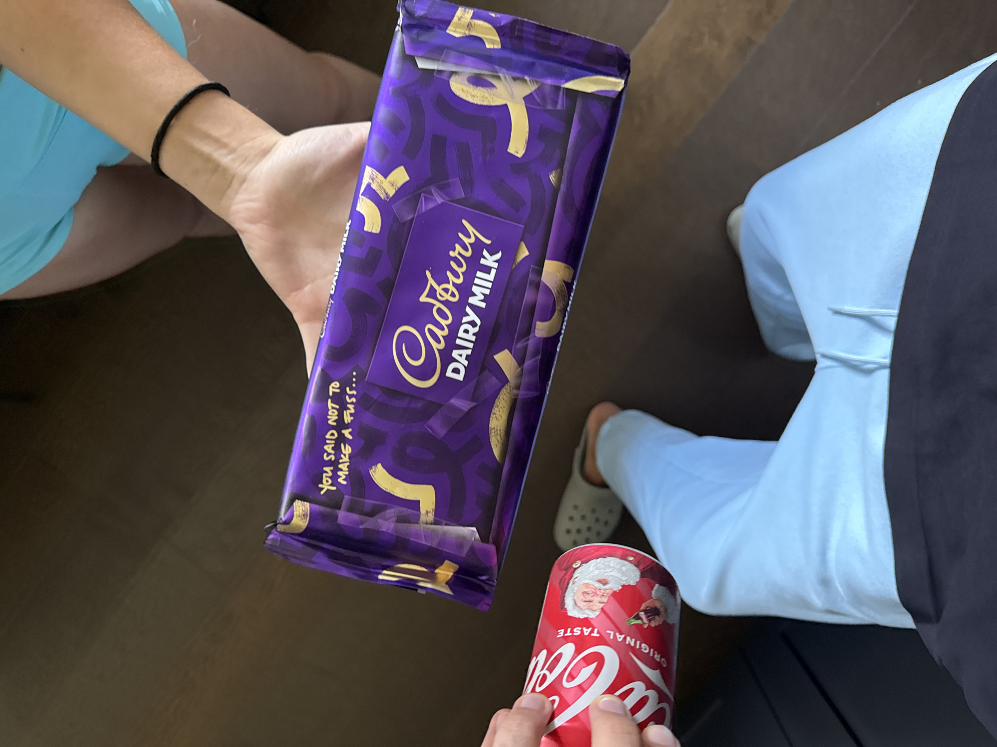This is a medium shot of a person's arms and legs as they hold a large Cadbury Dairy Milk chocolate bar and a can of Coca-Cola. The chocolate bar is purple with gold accents and has the words "Cadbury DAIRY MILK" printed on it. There is also text on the wrapper that reads "YOU SAID NOT TO MAKE A FUSS...". The Coca-Cola can is red with the iconic Coca-Cola logo and a picture of Santa Claus. The person is wearing light blue shorts and a black bracelet on their left wrist. Another person's legs, wearing light blue pants and grey clogs, are visible in the background. The setting appears to be indoors, with a dark wooden floor. The lighting suggests it could be daytime, but no specific time of day or weather conditions are discernible. The overall mood seems casual and relaxed.