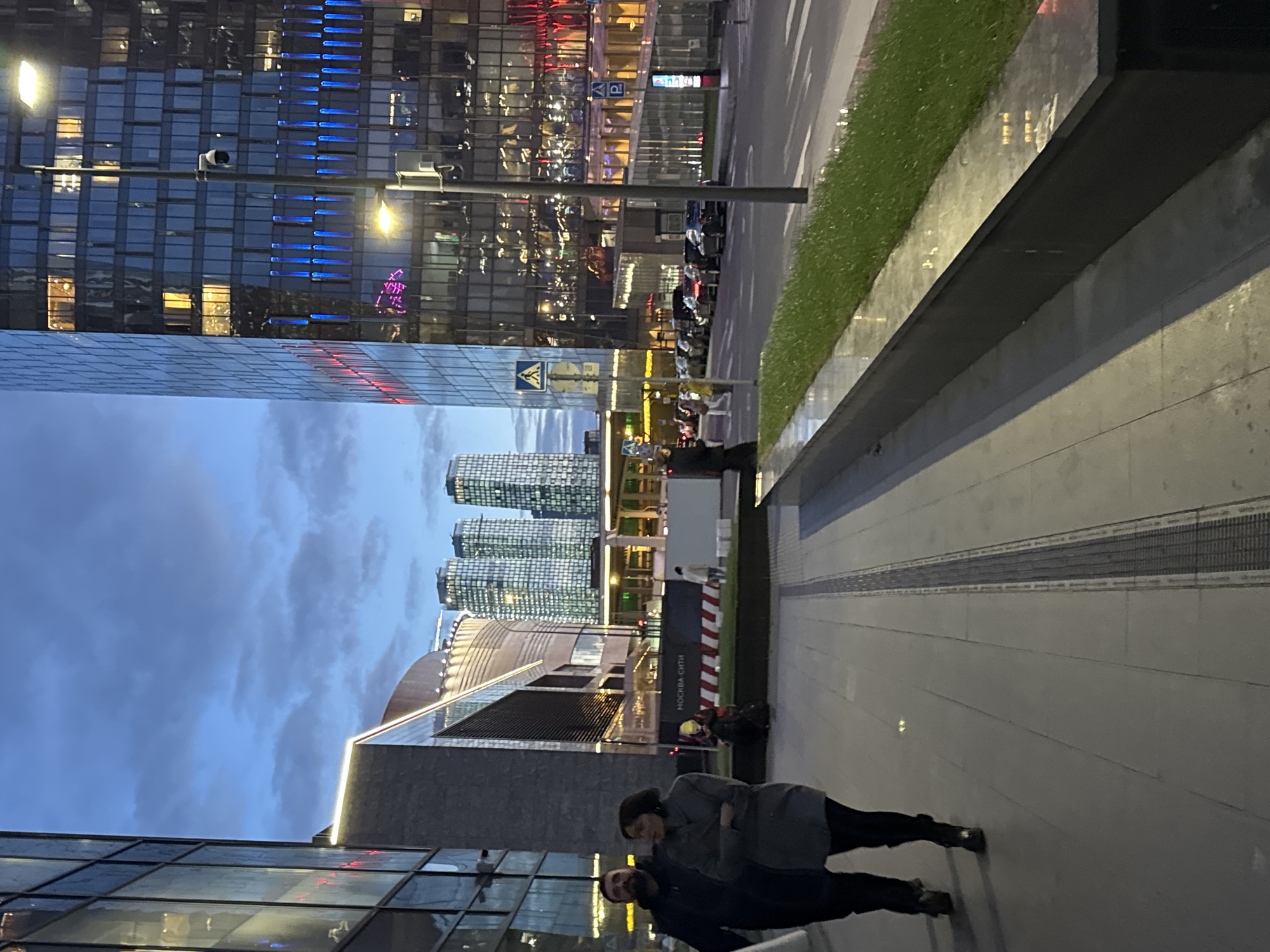 The image captures an urban street scene at dusk or early evening in Khimki, Russia, featuring modern architecture and a few pedestrians. The sky is a deep blue-grey with visible clouds, and artificial lights are illuminated throughout the scene.
In the foreground, a wide, light-colored paved sidewalk stretches across the bottom and right side of the frame. Two individuals are standing on this sidewalk. One, likely a woman, is wearing a dark coat and has her arms crossed, looking slightly to the left. The other, closer to the viewer, appears to be a man in a dark jacket, looking towards the first person. Their expressions are not distinctly visible.
To the left and in the background, numerous tall, glass-fronted skyscrapers dominate the view. Many windows are illuminated, and some buildings feature decorative blue and red lighting. A surveillance camera is mounted on one of these buildings in the upper left. Further down the street, on another building facade, faint, stylized text is visible, possibly "КАФЕ" (Cafe) and other similar establishment names.
A strip of neatly maintained green grass separates the sidewalk from a street or road, where several vehicles, including what appear to be parked cars or scooters, are visible. Streetlights illuminate the area. A blue pedestrian crossing sign (white figure on blue square) and a blue parking sign (white 'P' on blue square) are visible along the street.
In the mid-ground, further down the street, three distinct cylindrical high-rise buildings stand out against the sky. A white and red striped barrier or sign is positioned near the pedestrian path, bearing the text "МОСКВА СИТИ" (Moscow City), indicating the proximity to or view of the Moscow International Business Center. The overall setting is that of a bustling, modern city environment.