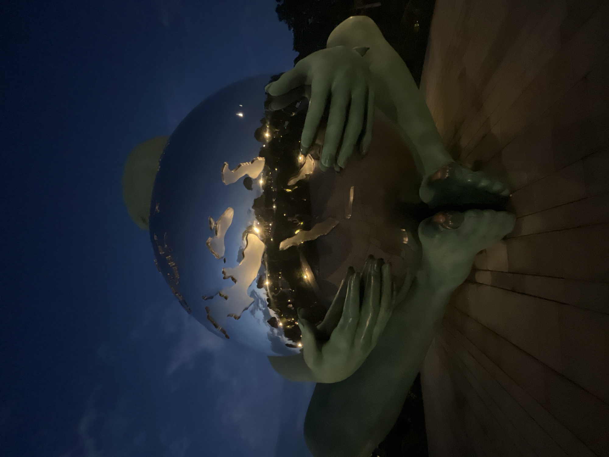 This media file is a low-angle shot of a sculpture of a person cradling a large, reflective globe at night. The sculpture, rendered in a pale green hue, is positioned in front of a wooden structure, possibly a wall or platform. The globe reflects the surrounding lights, including what appear to be city lights, and the dark blue night sky with scattered clouds.
The sculpture depicts a person with their legs bent and their feet visible near the bottom of the frame. Their hands are wrapped around the globe, which seems to represent the Earth, showing landmasses and bodies of water. The reflection on the globe is clear enough to discern continents, likely Europe and Africa. The overall mood is serene and contemplative, with the lighting emphasizing the reflective surface of the globe and the dark night sky.
The time of day is clearly night, indicated by the dark sky and the artificial lights reflected on the globe. There are no visible weather cues beyond the presence of clouds. Location cues are limited to the reflective representation of Earth and the presence of artificial lights, suggesting an urban or developed area. No text is visible in the image. The context provided places this scene in Krasnodar, Russia.
