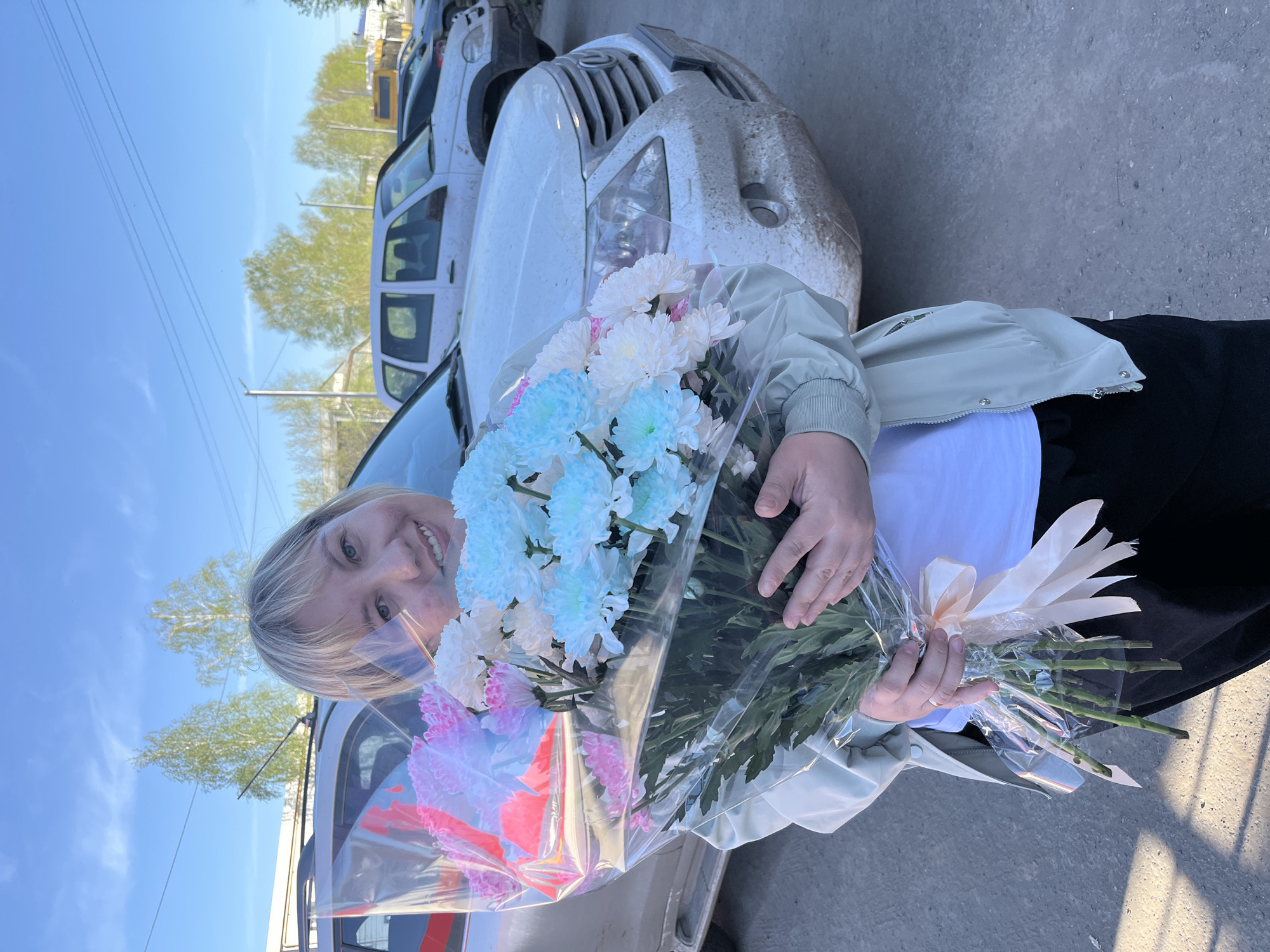 A woman is holding a bouquet of flowers wrapped in clear plastic. The bouquet contains light blue, white, and light pink flowers. She's wearing a light green jacket over a white top and dark pants. In the background, a dirty white car is visible, along with other parked vehicles. The sky is blue, with trees and power lines in the distance. The location is outdoors on a paved surface. Based on visual cues, the city is Izhevsk, Russia.